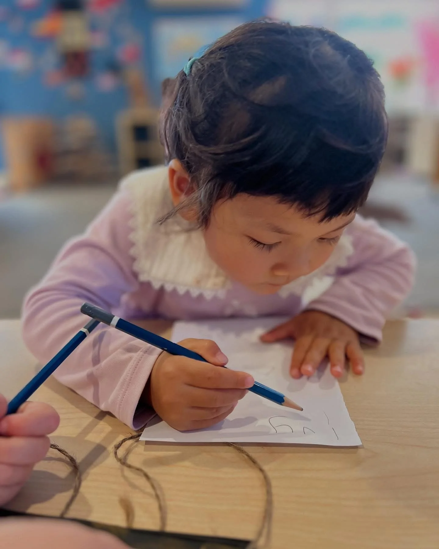 A young girl with short black hair and a blue hair clip is drawing with a blue pencil on a piece of white paper on a wooden table.