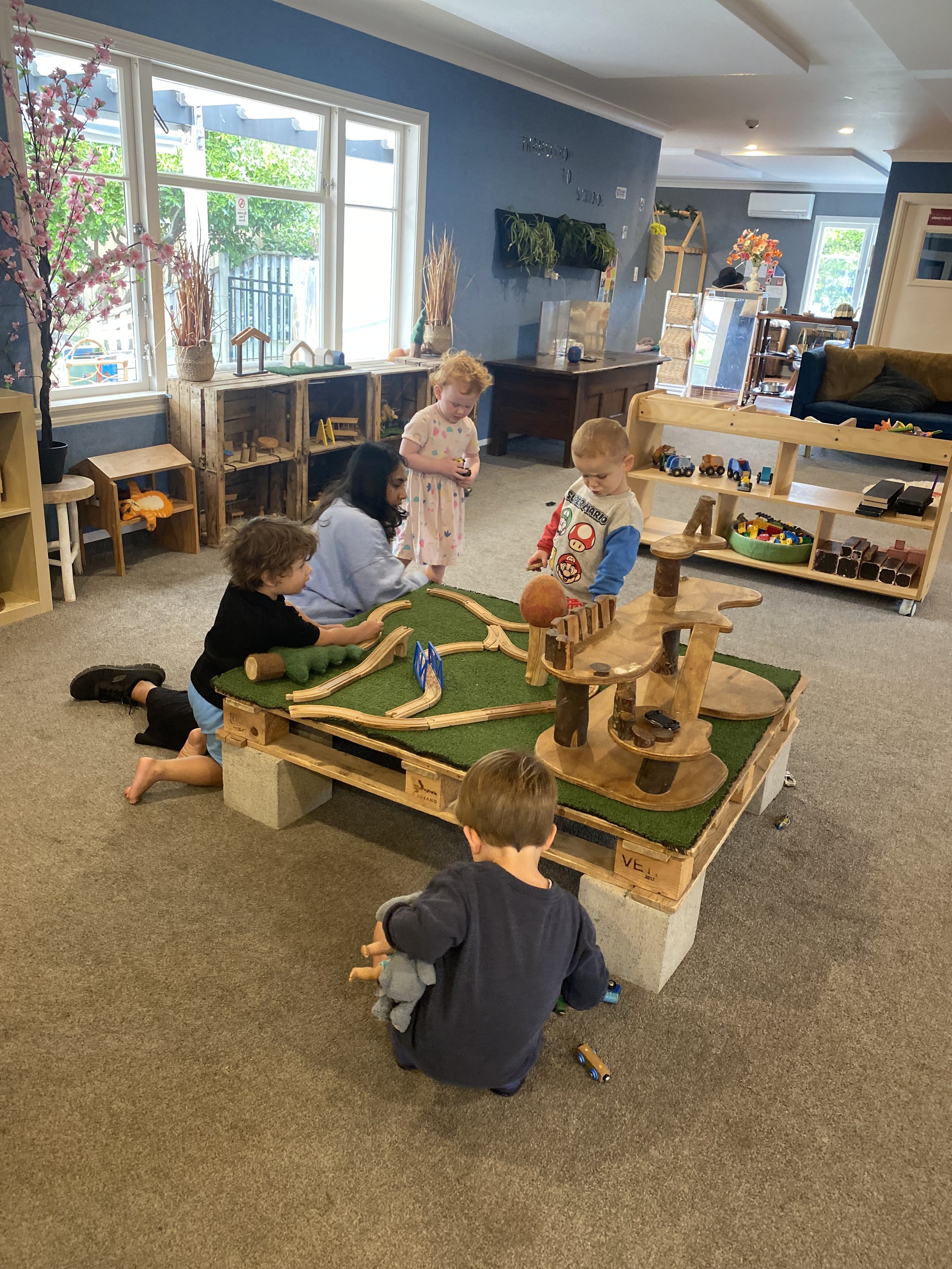 Children playing with a wooden train set in a brightly lit indoor playroom.