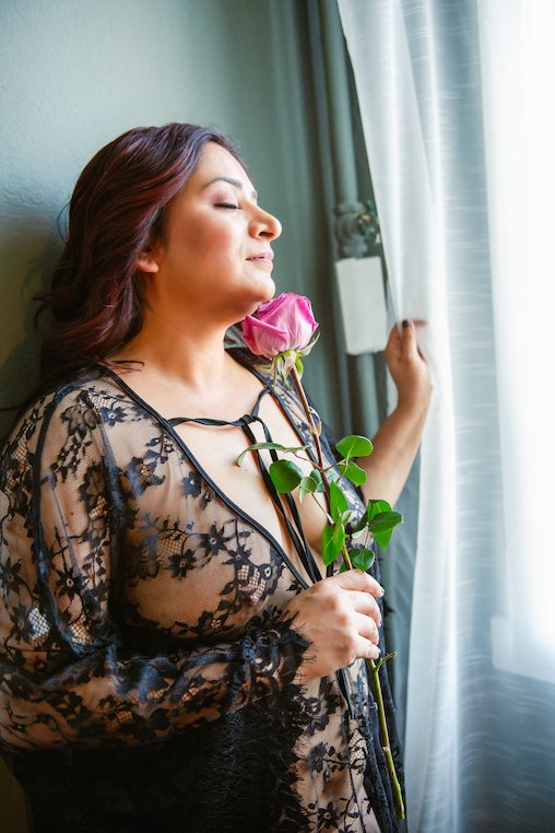 Person in a lace robe holding a pink rose near a window.
