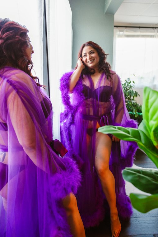 A woman in a purple sheer robe stands in front of a mirror, smiling. The setting appears to be a bright room with natural light and some greenery visible.