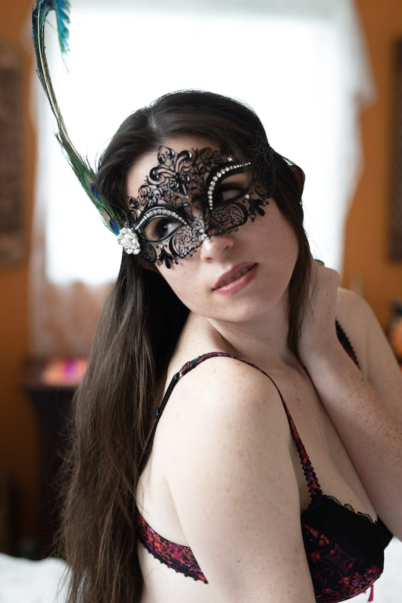 Person wearing a black lace masquerade mask with a peacock feather, dressed in a colorful bra, looking to the side.