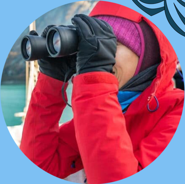 Person in a red winter jacket and purple knit hat looking through binoculars outdoors, with a body of water in the background.
