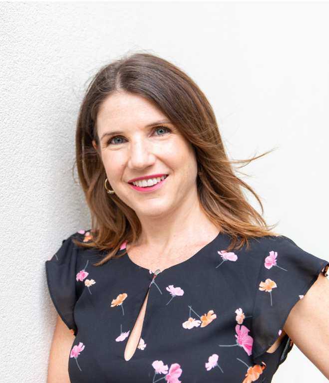 Smiling woman with brown hair and earrings leaning against a light-colored wall, wearing a black floral dress.