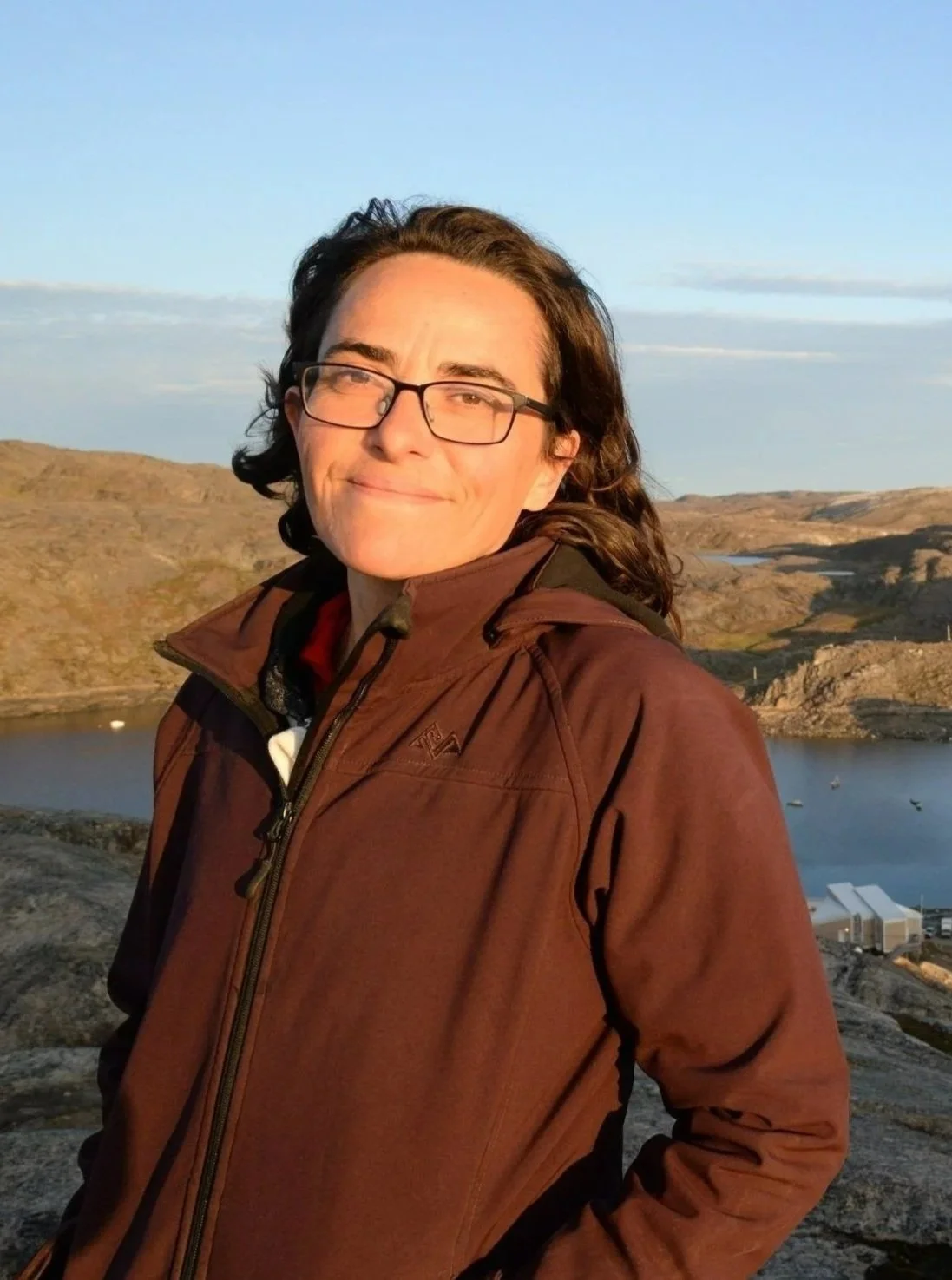 A woman with long curly hair, glasses, and a brown jacket standing outdoors near a body of water with hills in the background, during sunset or sunrise.