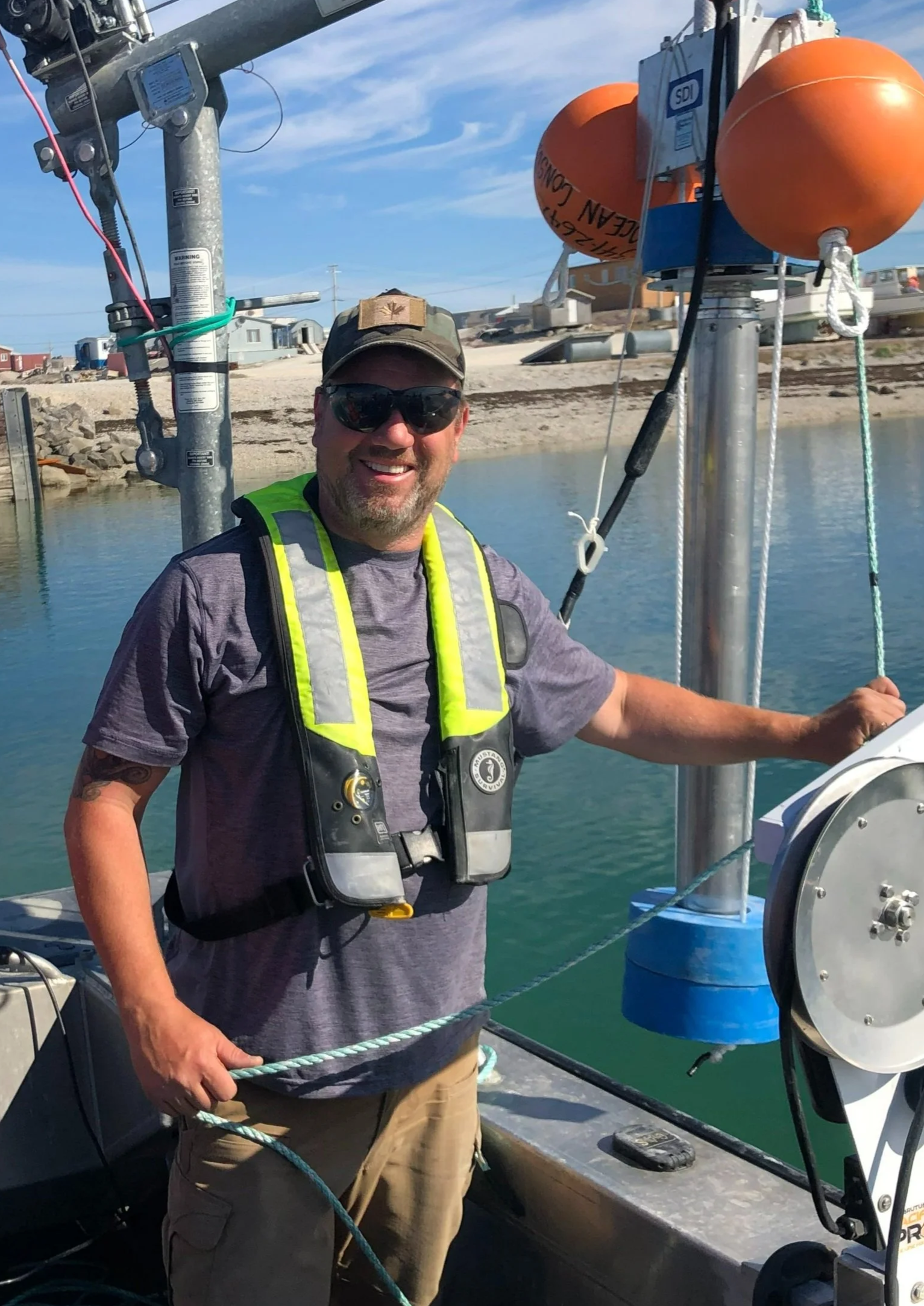 A smiling man wearing a gray t-shirt, sunglasses, a life vest, a cap, and khaki shorts, standing on a boat near water with a shoreline and houses in the background.