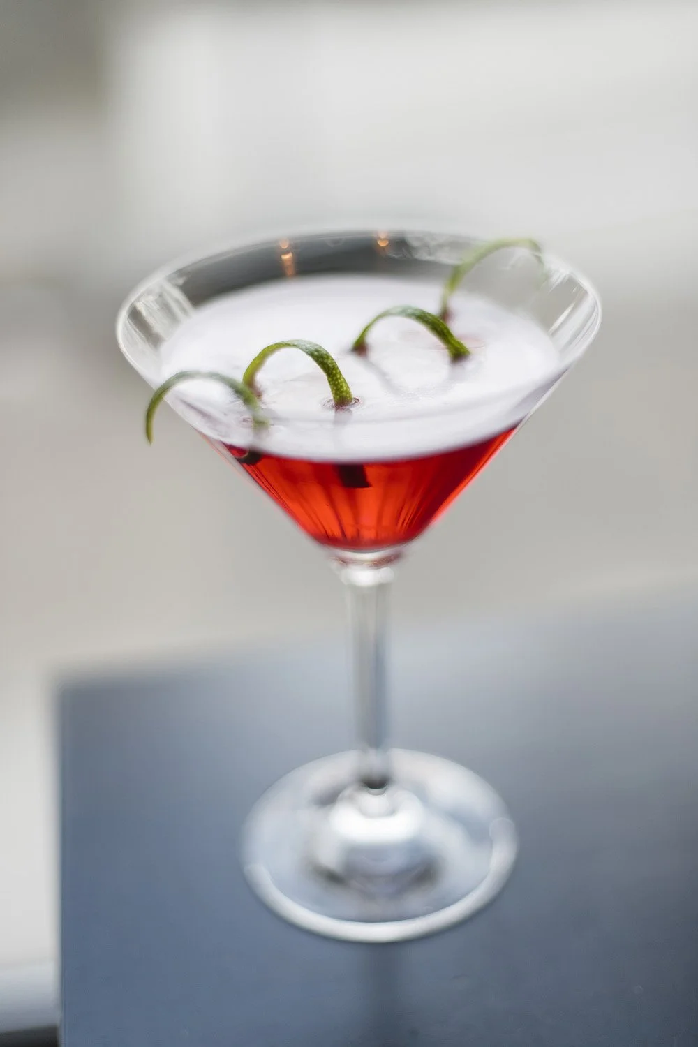 Rød cocktail servert i martiniglass med skum og limezest.