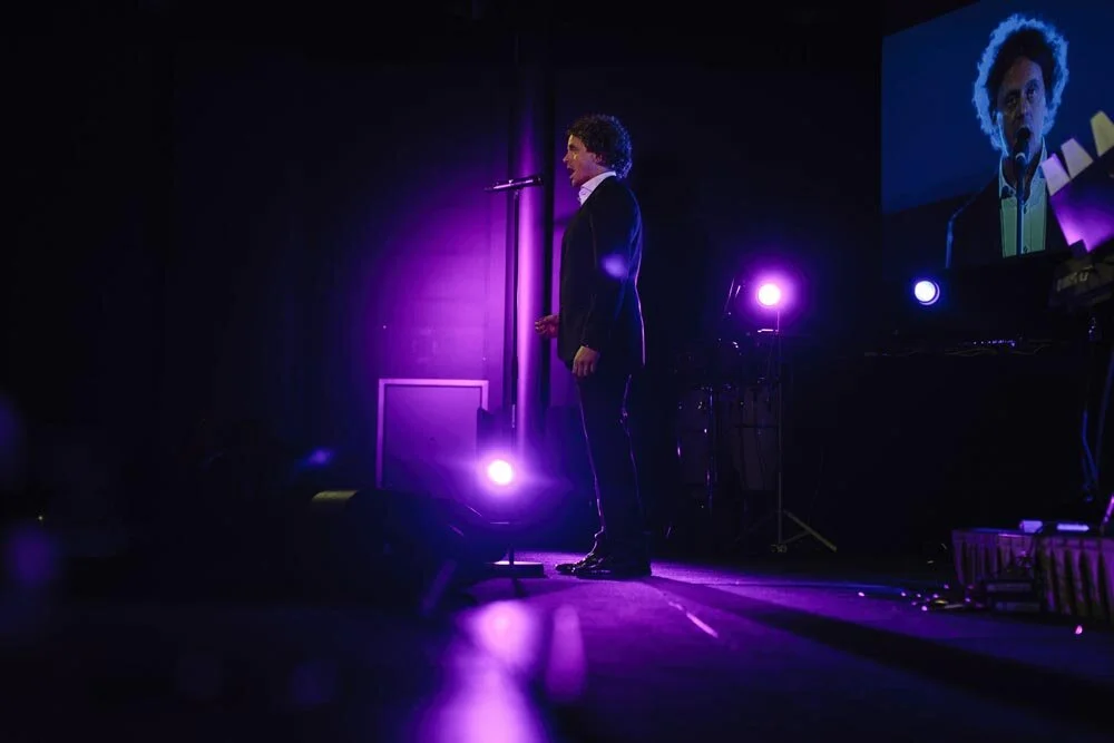 A man in a suit standing on stage with a microphone and purple lighting, with a large screen displaying his image in the background.