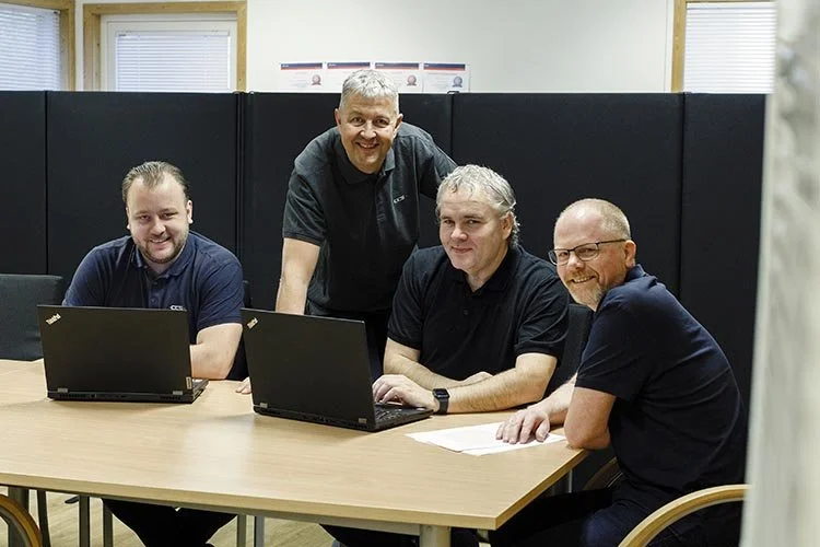 Group of four men sitting around a table with laptops, discussing or working together in an office setting.