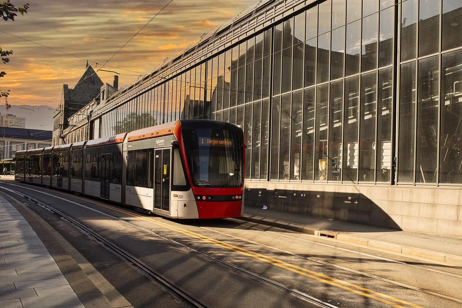 Tram in Bergen city reflecting sunset, adjacent to glass building.