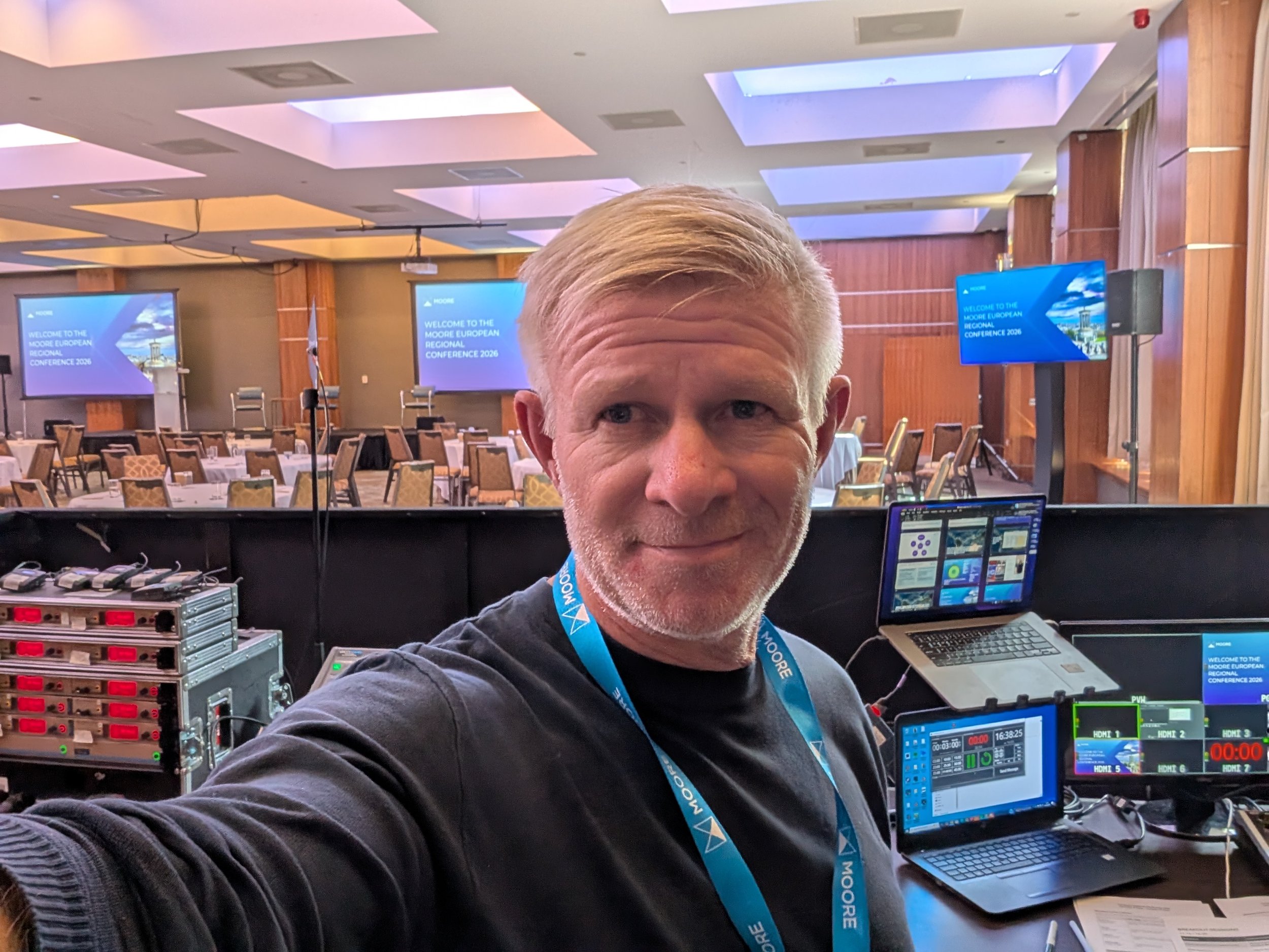 A man with gray hair and a mustache looking at the camera, wearing a black shirt and a blue conference badge, in a conference room with tables, chairs, and large screens displaying the text 'Welcome to the Moore European Regional Conference 2026.'
