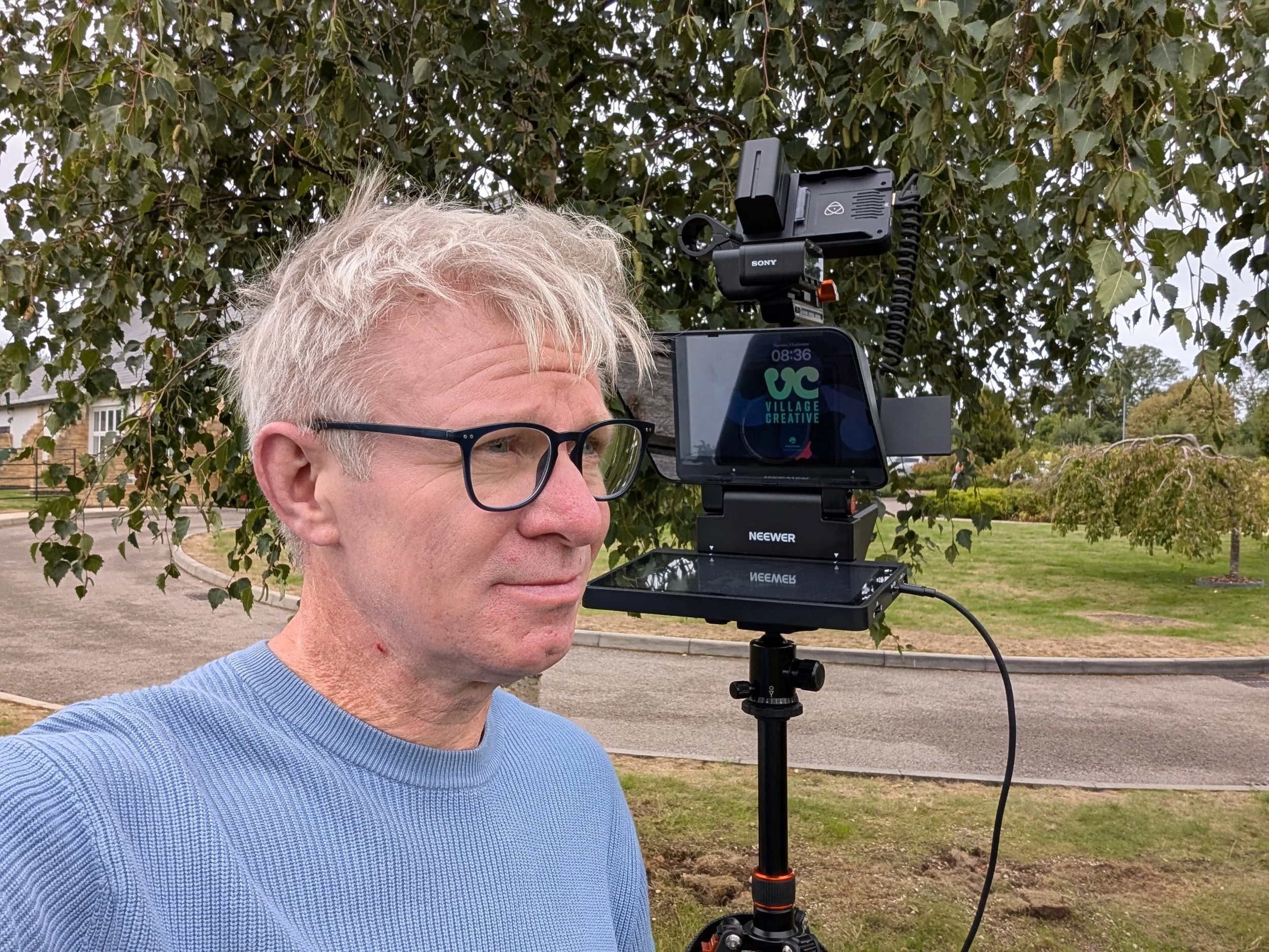 A man with gray hair and glasses operating video equipment outdoors, with trees and houses in the background.
