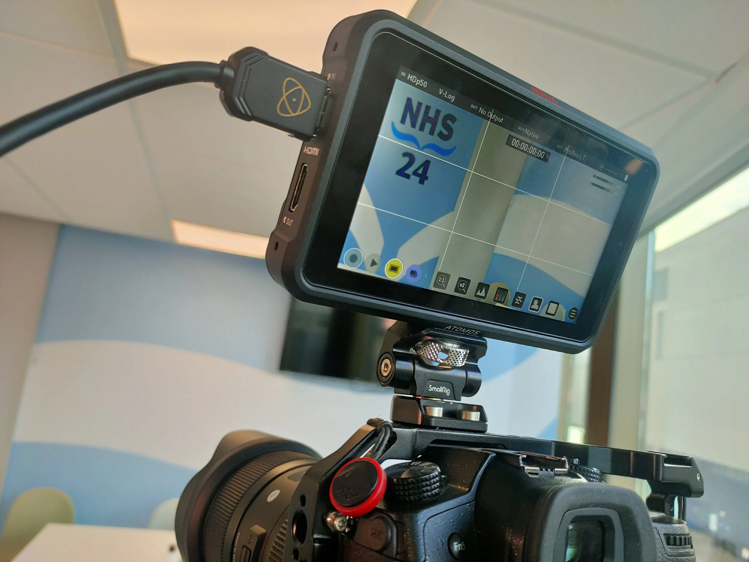 A camera mounted on a stabilizer captures a shot of a monitor displaying the NHS logo, with a cable plugged into the camera.