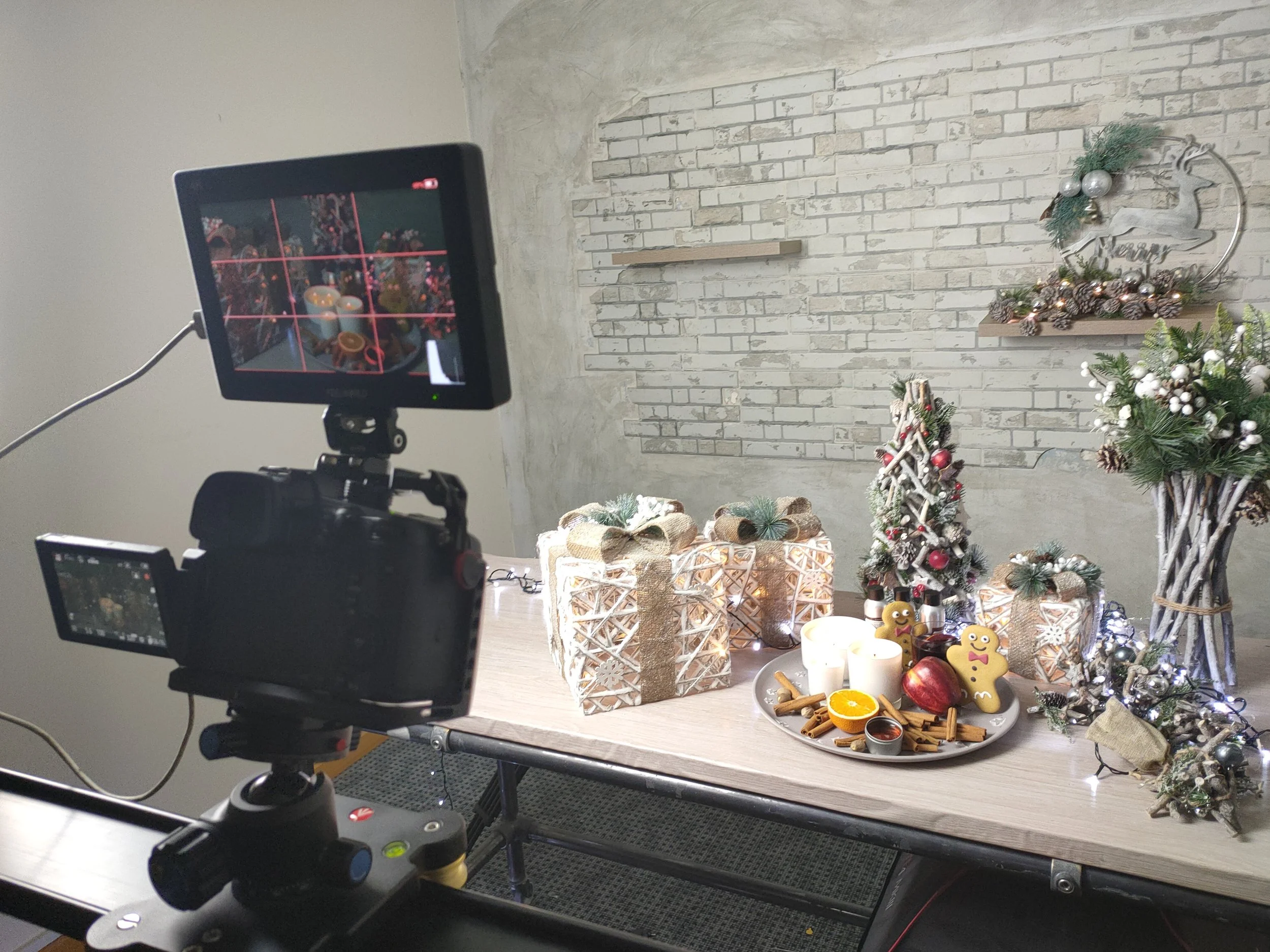 A camera on a tripod capturing a Christmas-themed display with holiday decorations, wrapped gifts, candles, and festive ornaments against a white brick wall.