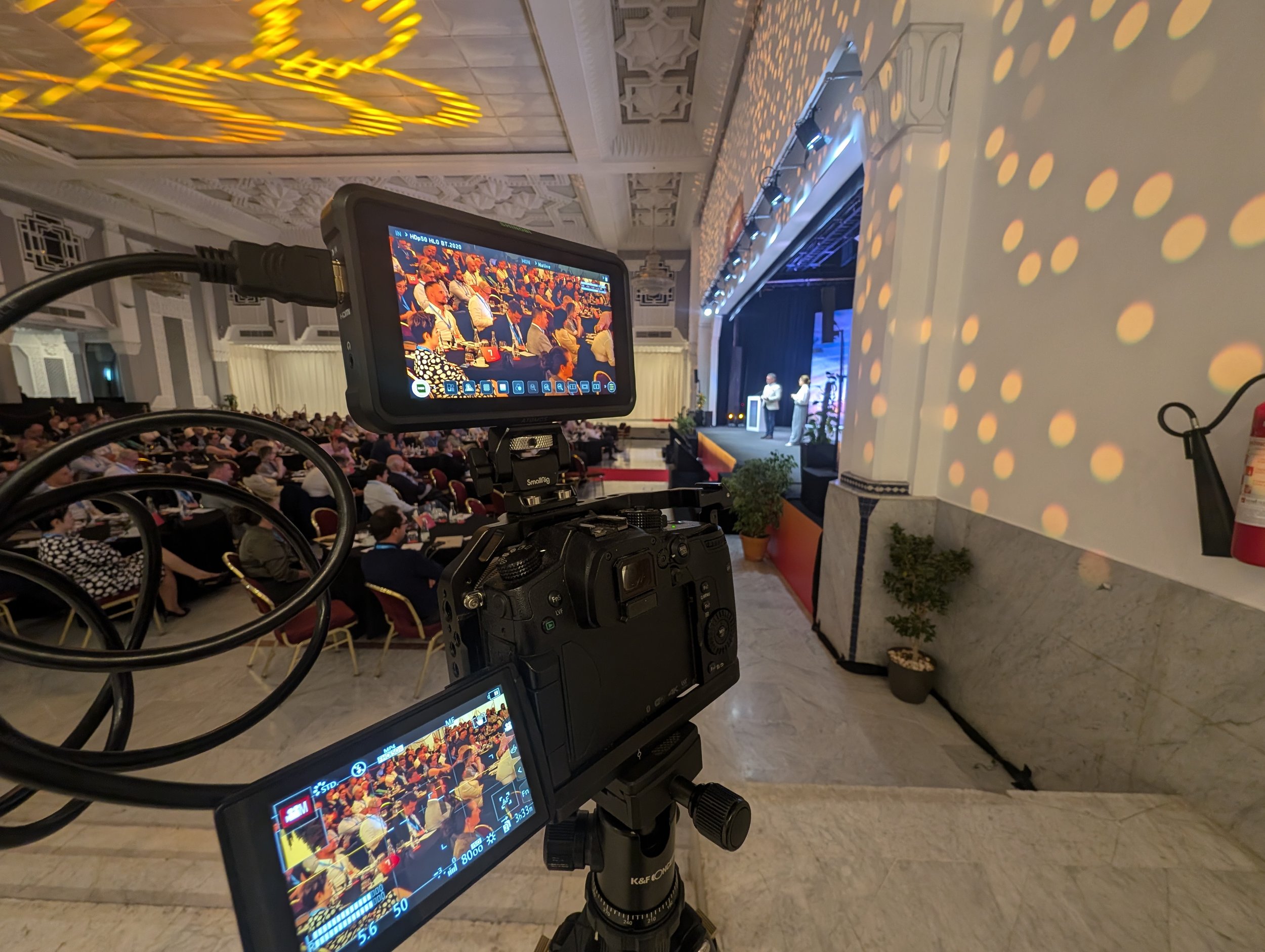 A camera on a tripod recording an indoor conference or presentation with an audience seated at tables. Two people are on stage speaking, with decorative lighting and intricate architectural details in the background.