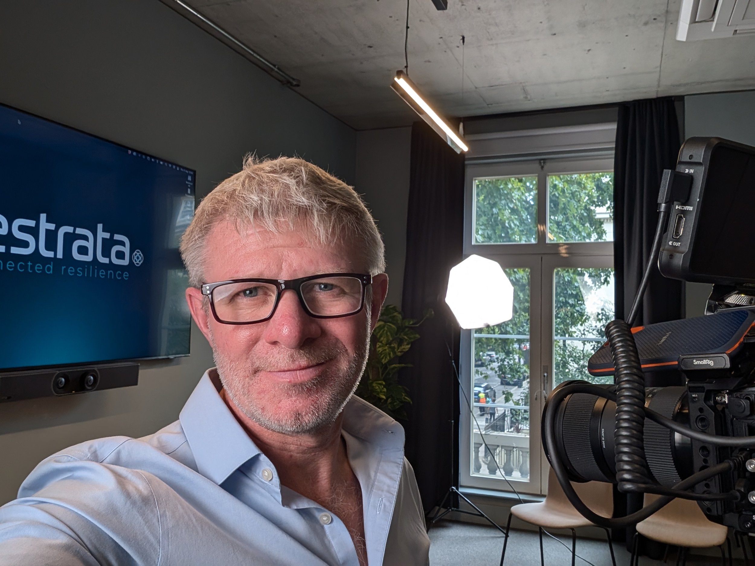 A man with glasses and light-colored hair taking a selfie in an office or studio setting. Behind him is a large monitor with a screen displaying the words 'e. strato' and a logo, along with professional lighting equipment and a window showing green trees outside.