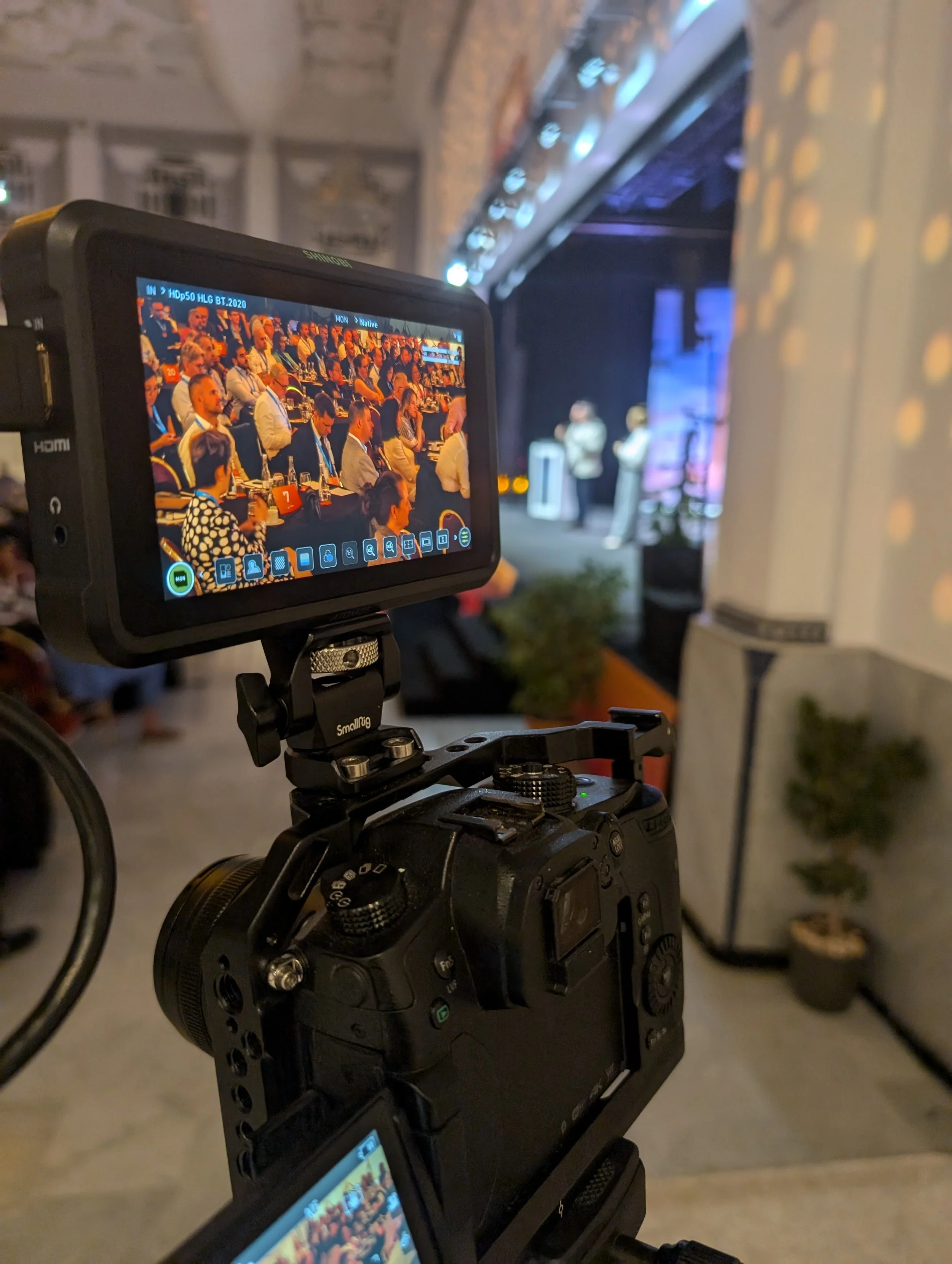 A professional camera on a tripod recording a conference or presentation on stage, with an audience seated at tables in a well-lit room.