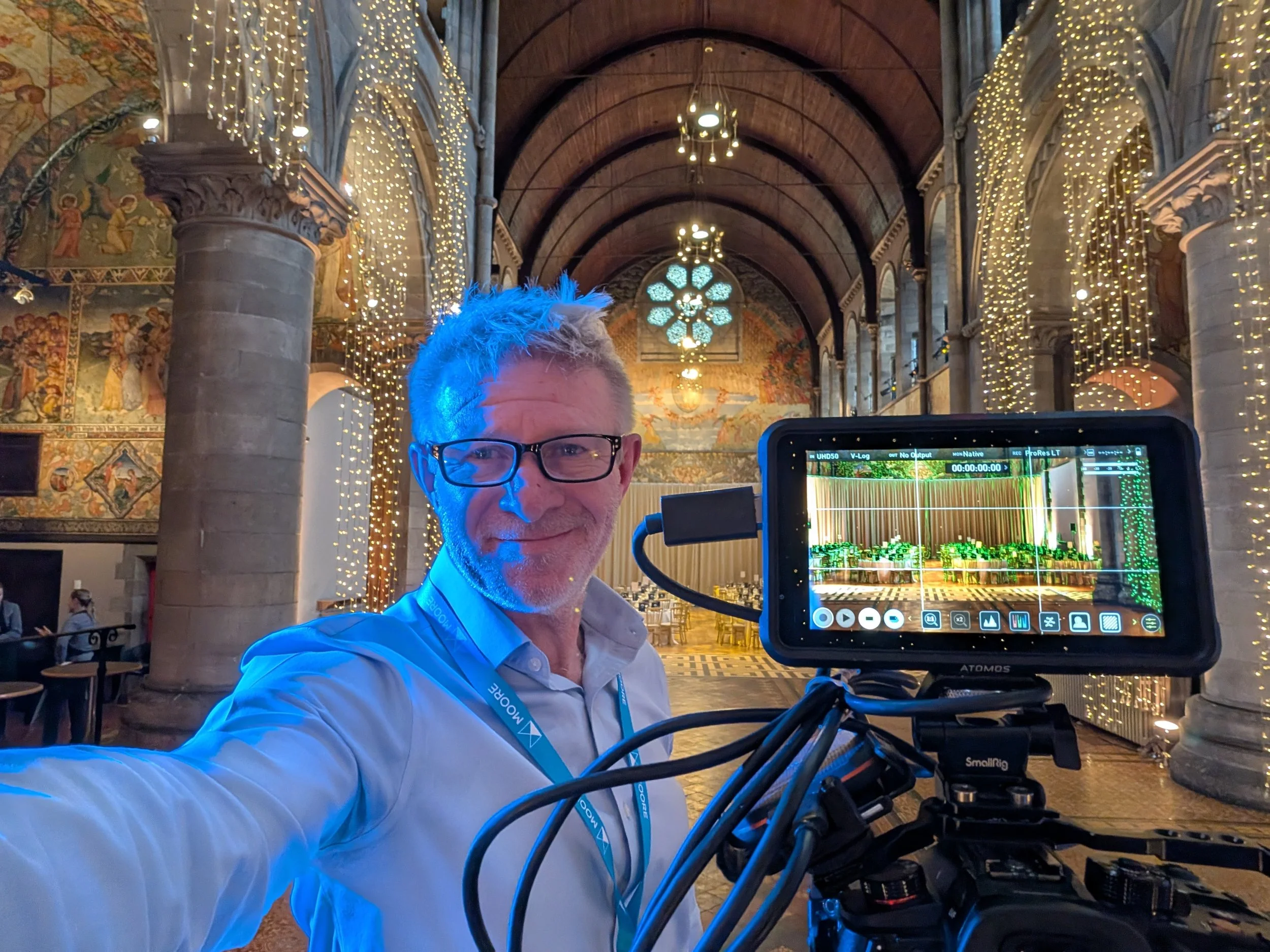 A person with glasses and light-colored hair is taking a selfie inside a decorated church or cathedral with fairy lights hanging from the ceiling and murals on the walls. The person is smiling and is in front of a video camera monitor.