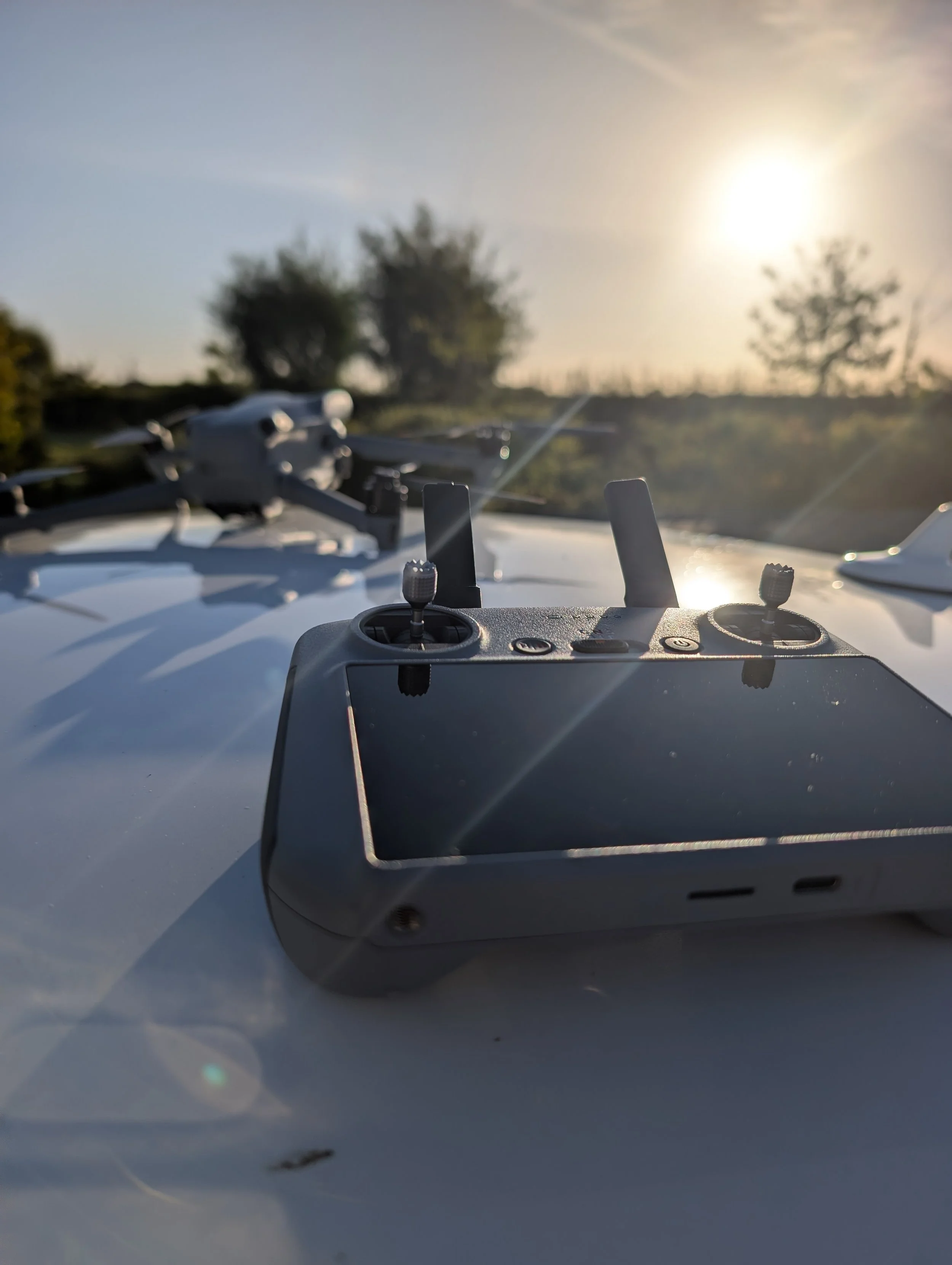 Close-up of a drone controller with a small drone on a white surface, outdoors with a sunlit sky and blurred trees in the background.