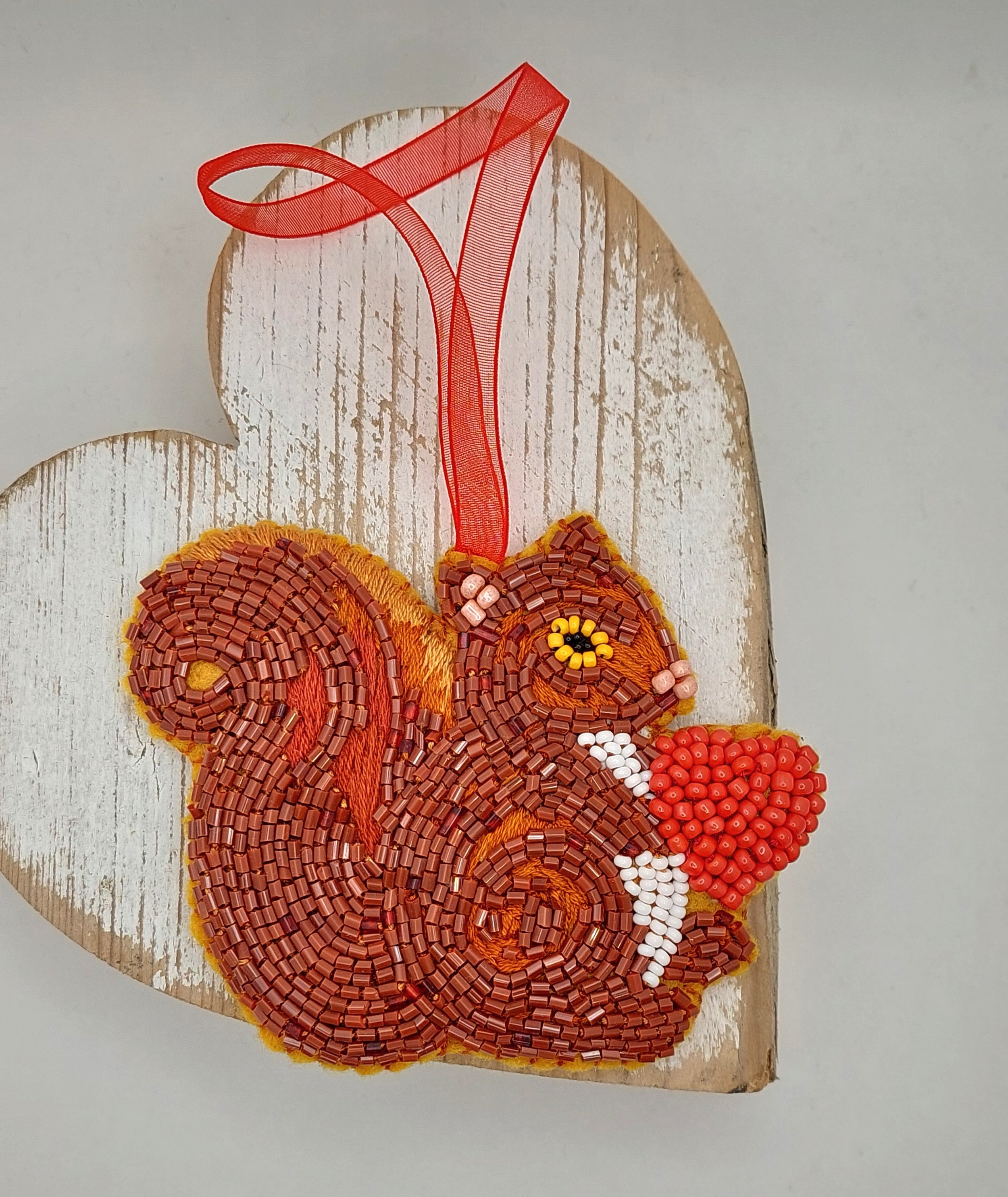 Hand-sewn red squirrel bead embroidery decoration holding a red heart, on felt with detailed beading and a red ribbon for hanging – 78mm x 95mm.