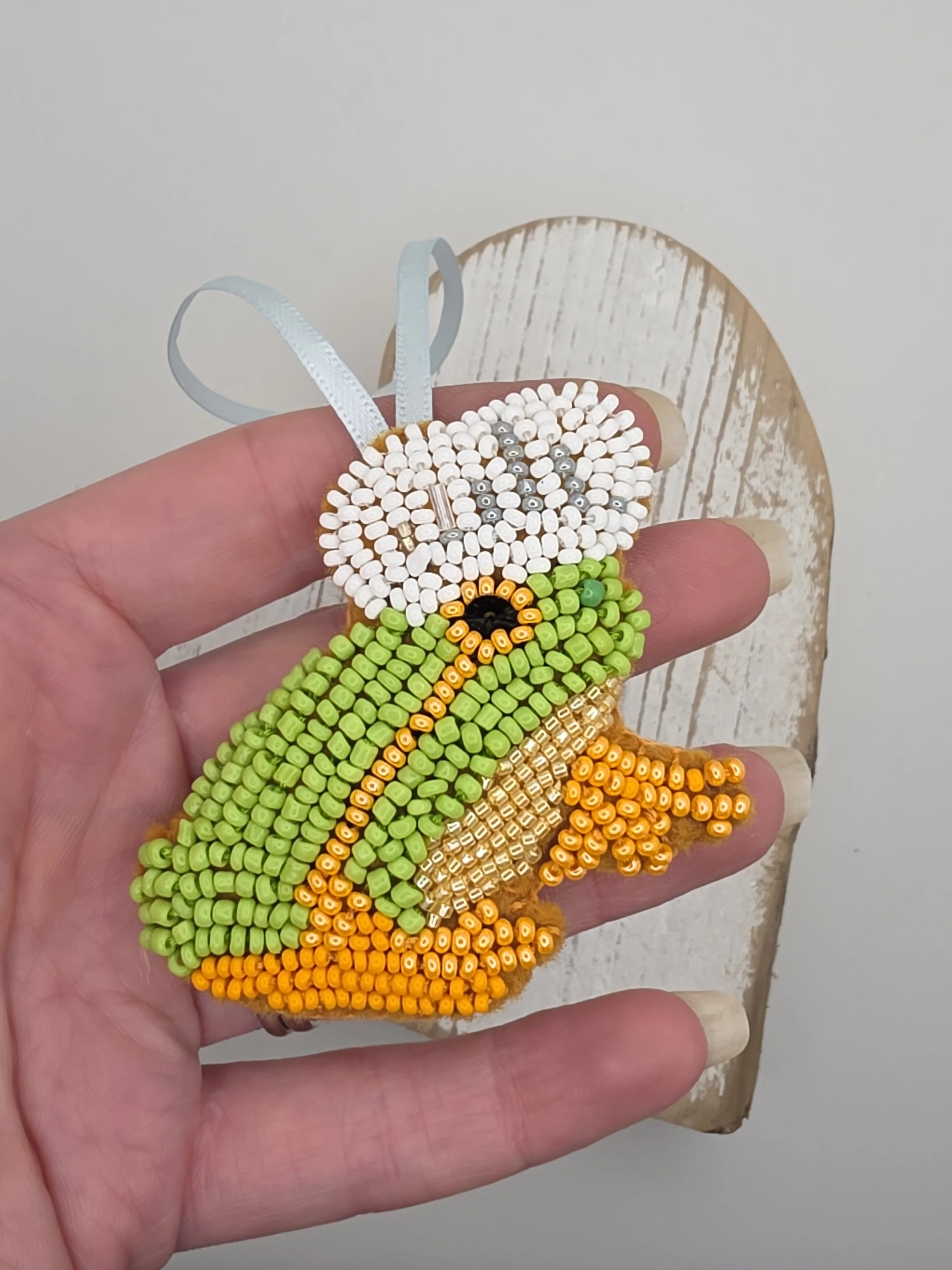 Hand holding a handmade beaded frog decoration with a white shower hat, green body, orange legs, and black sequin eye, embroidered on brown felt with pale blue hanging ribbon.