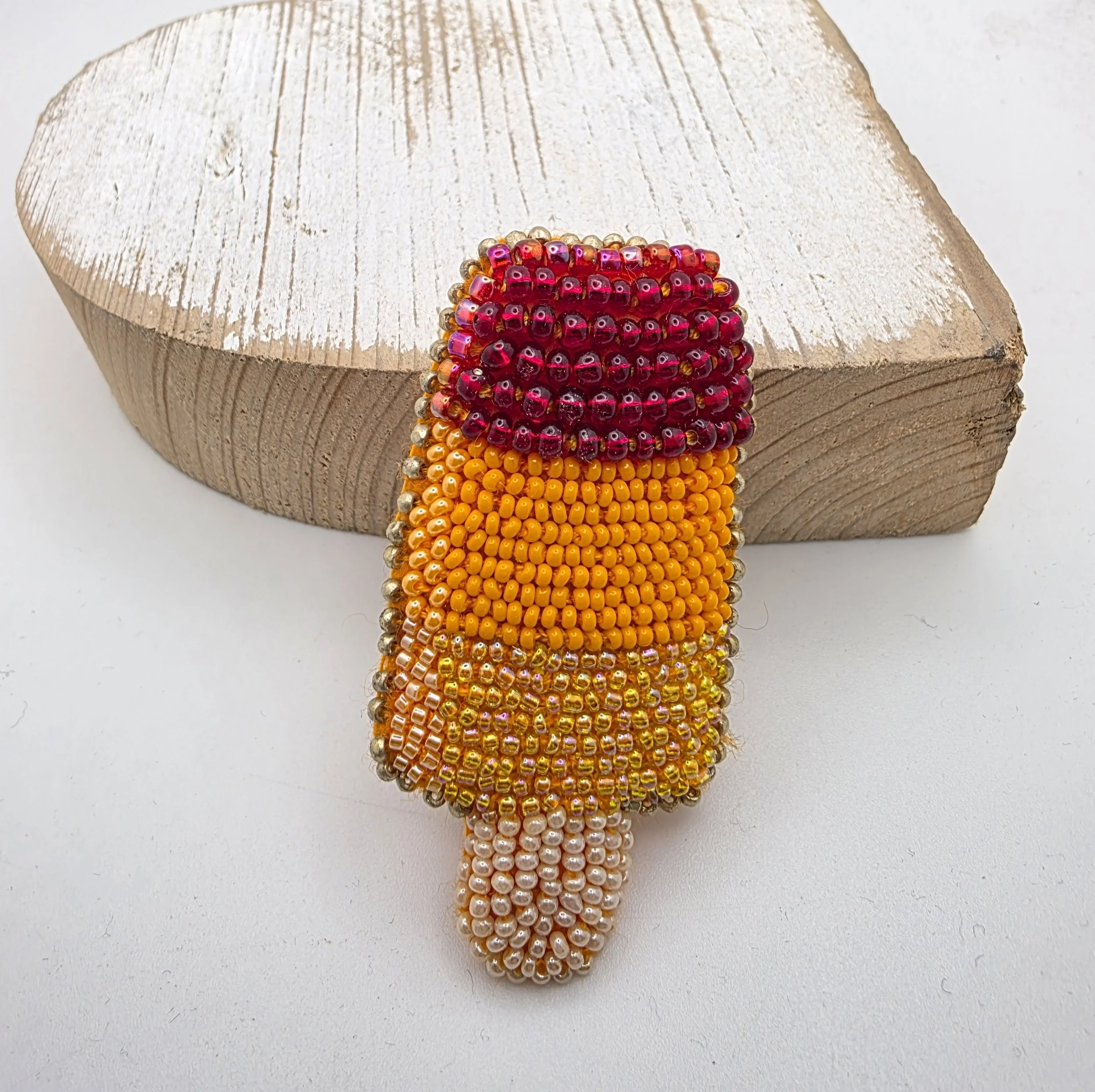 Handmade beaded ice lolly brooch leaning against distressed wood background. Features lemon, orange, and strawberry stripes with a beaded stick.