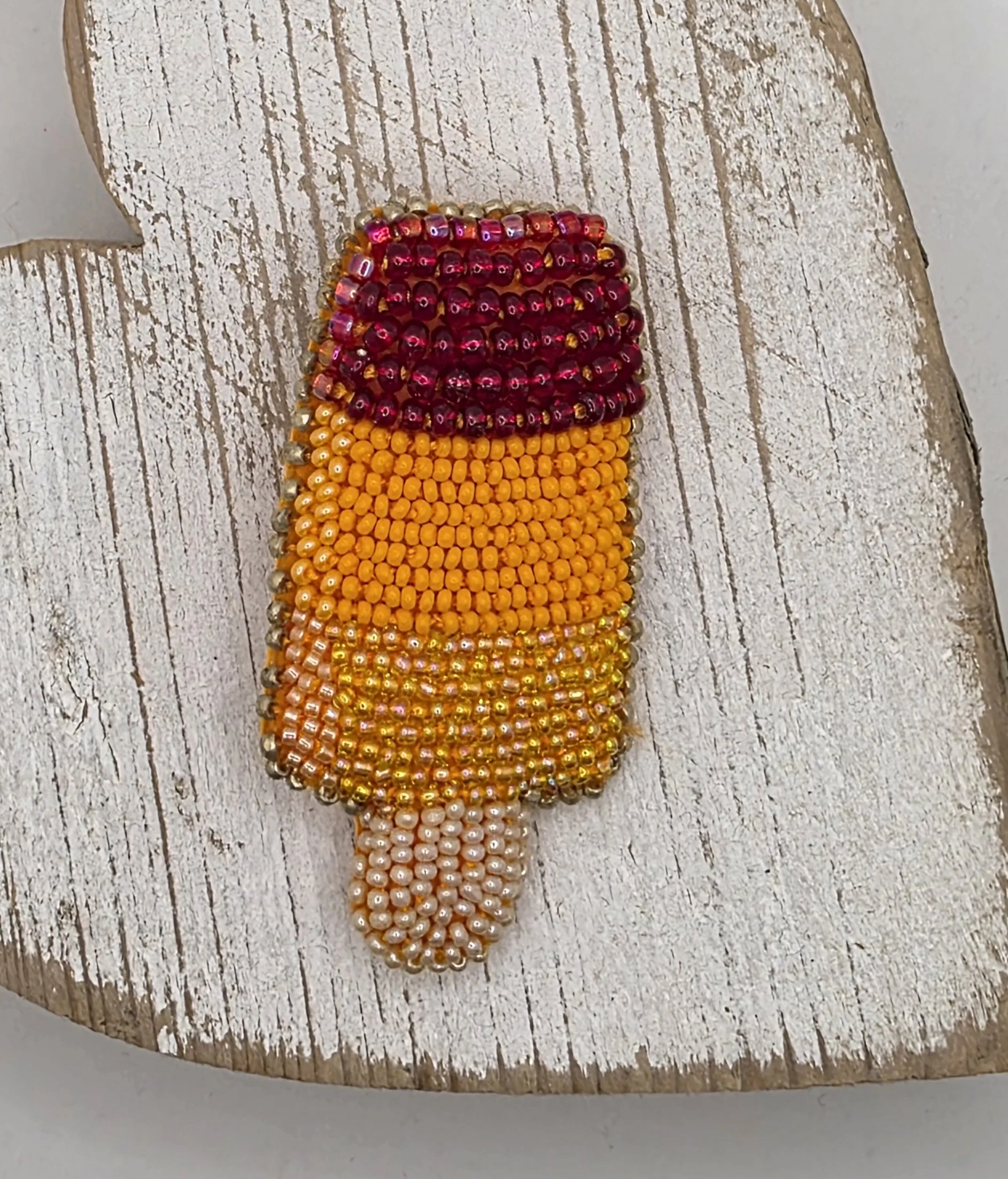 Close-up of embroidered ice lolly brooch laying flat on rustic wood. Shows detailed beadwork in yellow, orange, and pink with gold stitched edging.