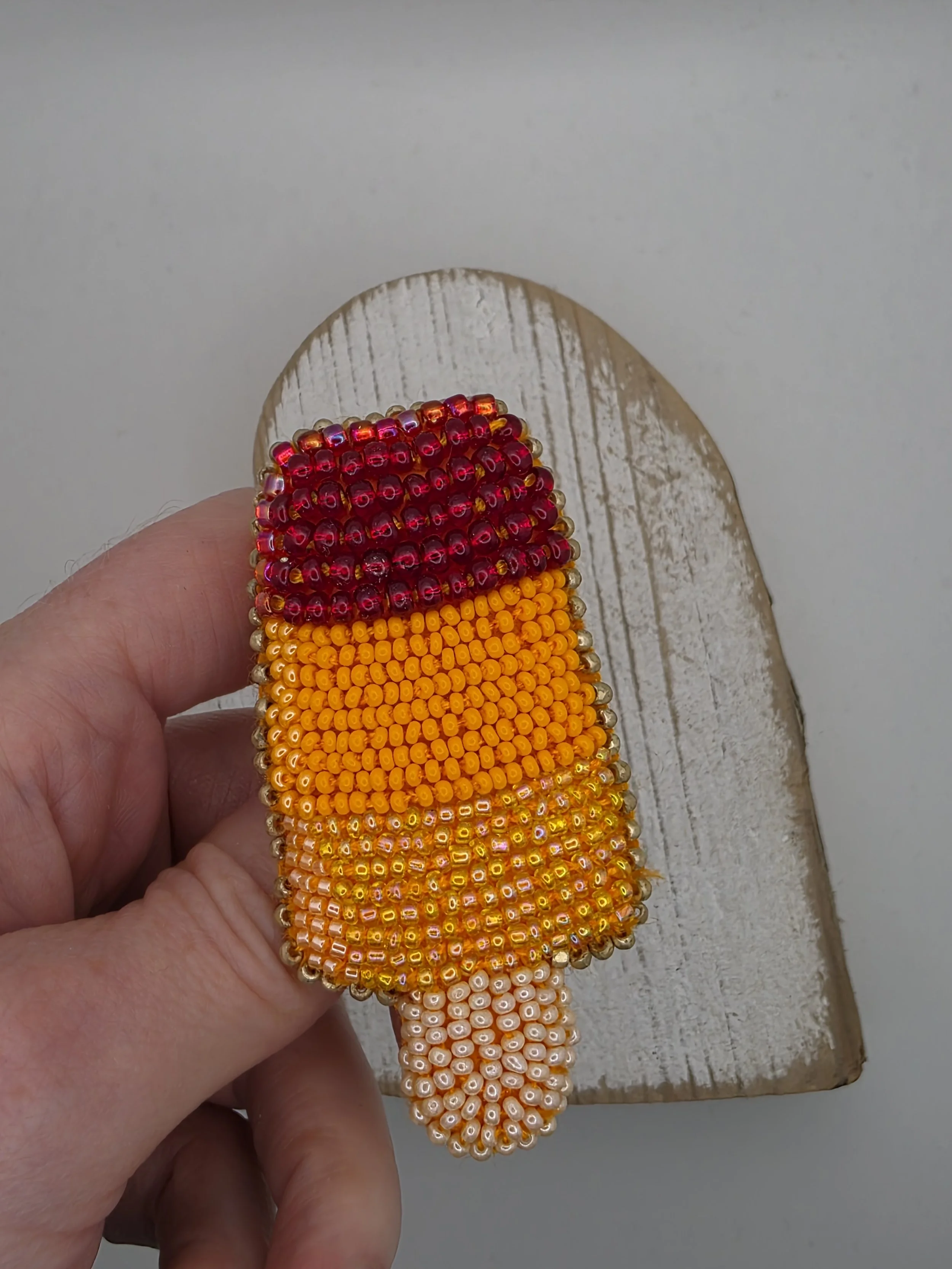 Colourful beaded ice lolly brooch held in hand to show scale. Features strawberry, orange, and lemon stripes with intricate hand-stitched beadwork.