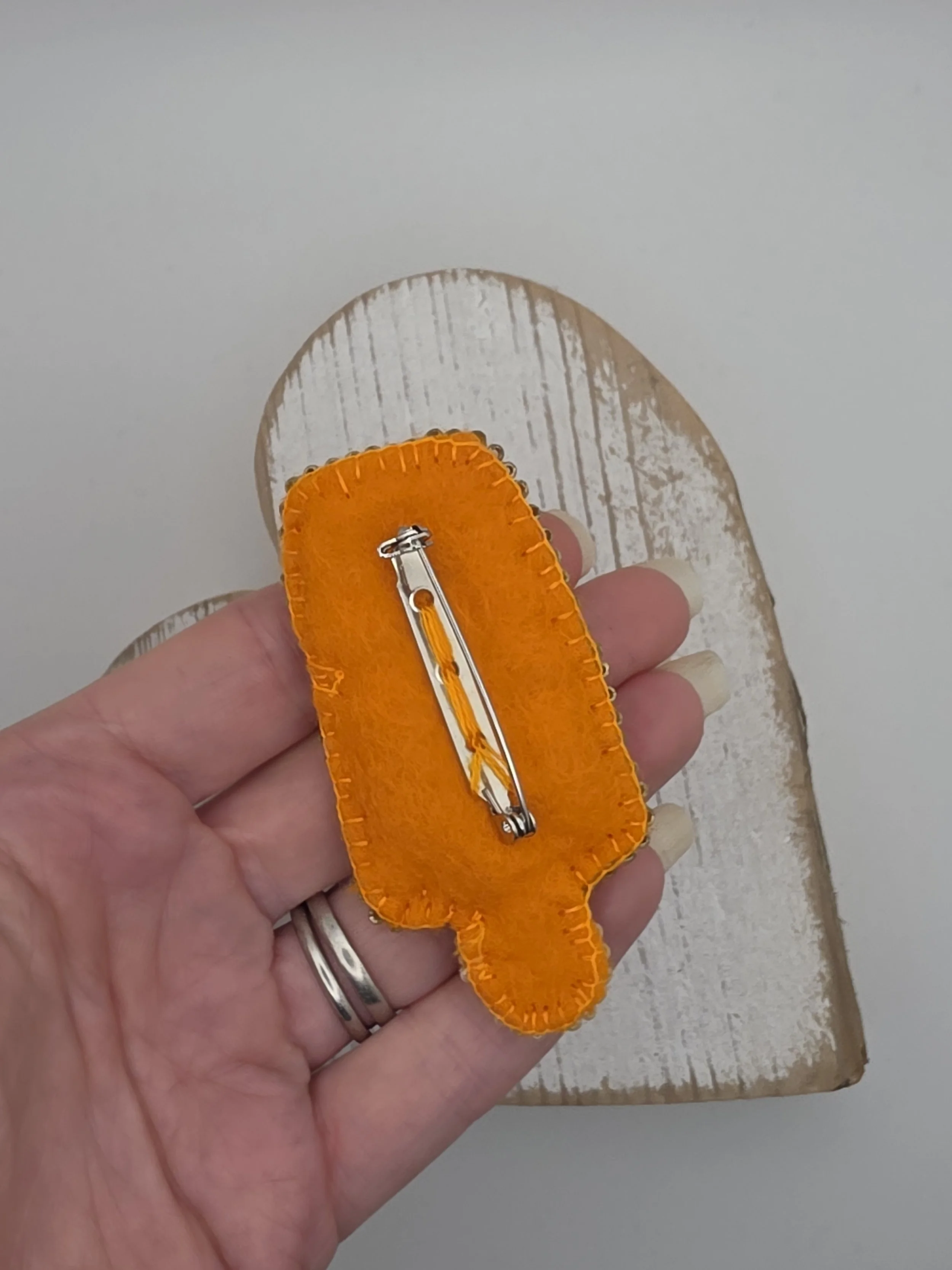 Reverse side of embroidered brooch showing orange felt backing and silver brooch pin securely stitched in place.