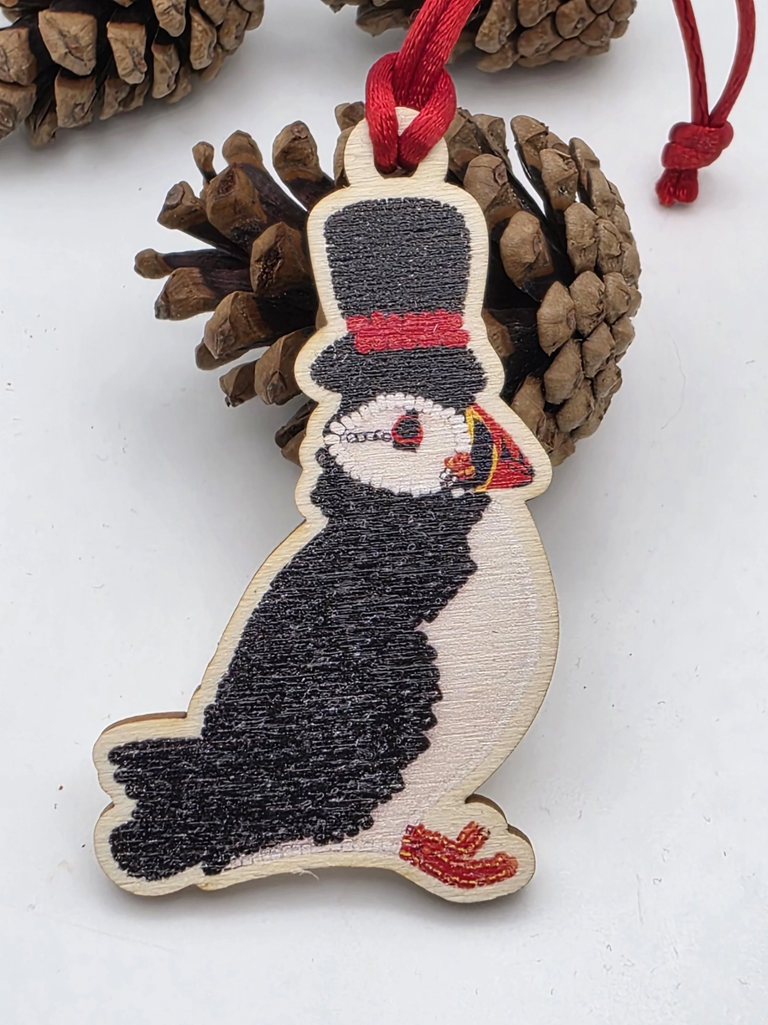 Wooden puffin ornament wearing a black top hat with red band, with a red satin cord, resting beside a small pine cone on a white background.