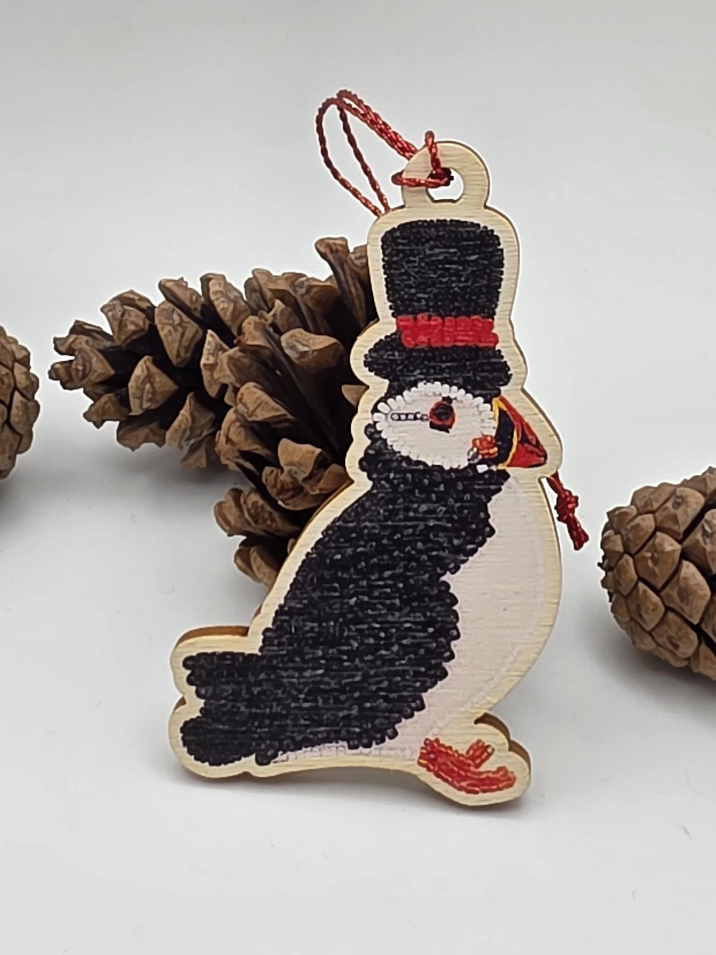 Wooden puffin Christmas ornament with red sparkly thread hanging and top hat, puffin decoration for bird lovers, styled beside a pine cone on white surface.