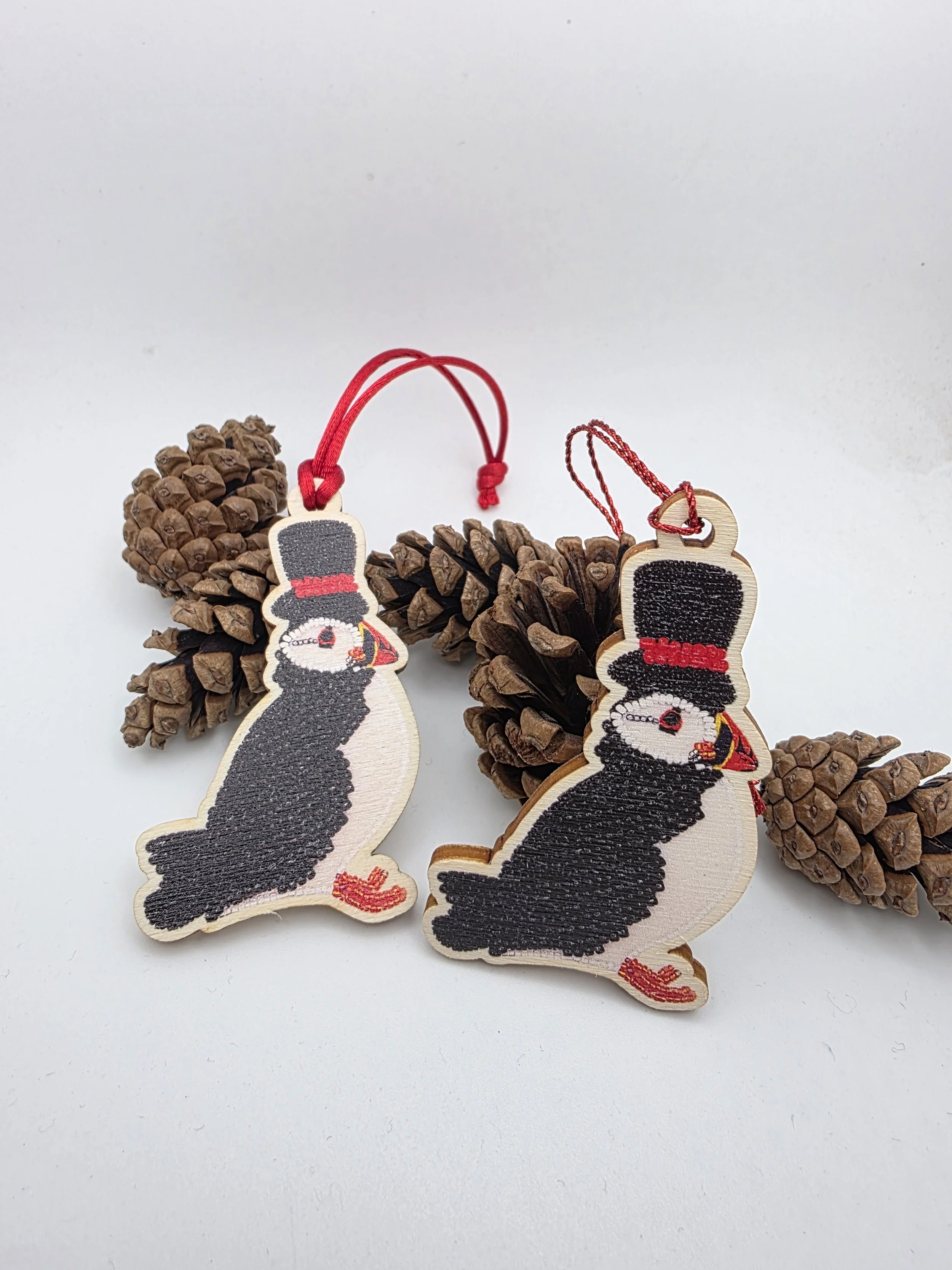 Quirky wooden puffin hanging decoration with red satin cord or sparkly thread, photographed beside some pine cones for festive coastal Christmas décor.