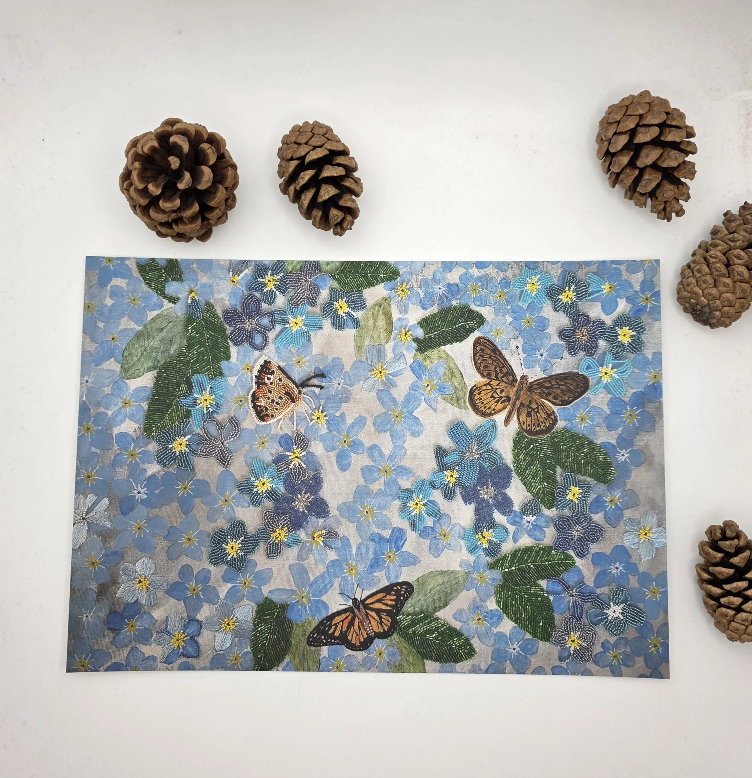 An open edition A5 Forget-Me-Not art print featuring blue flowers and three butterflies, photographed flatlay on a white background beside natural pinecones.