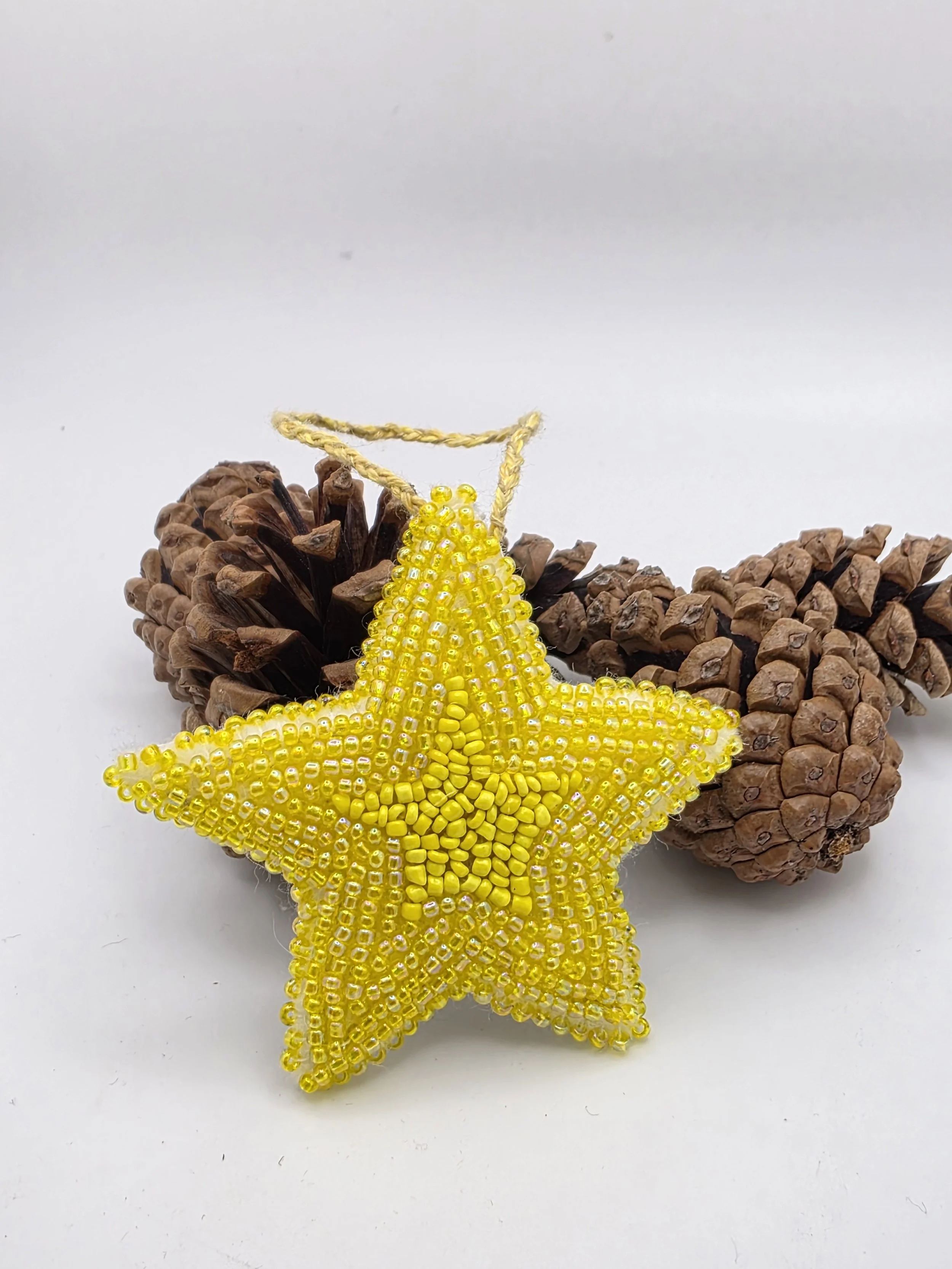Handmade yellow star decoration with intricate bead embroidery, displayed upright against rustic pine cones on a bright white background, ideal for home décor inspiration.