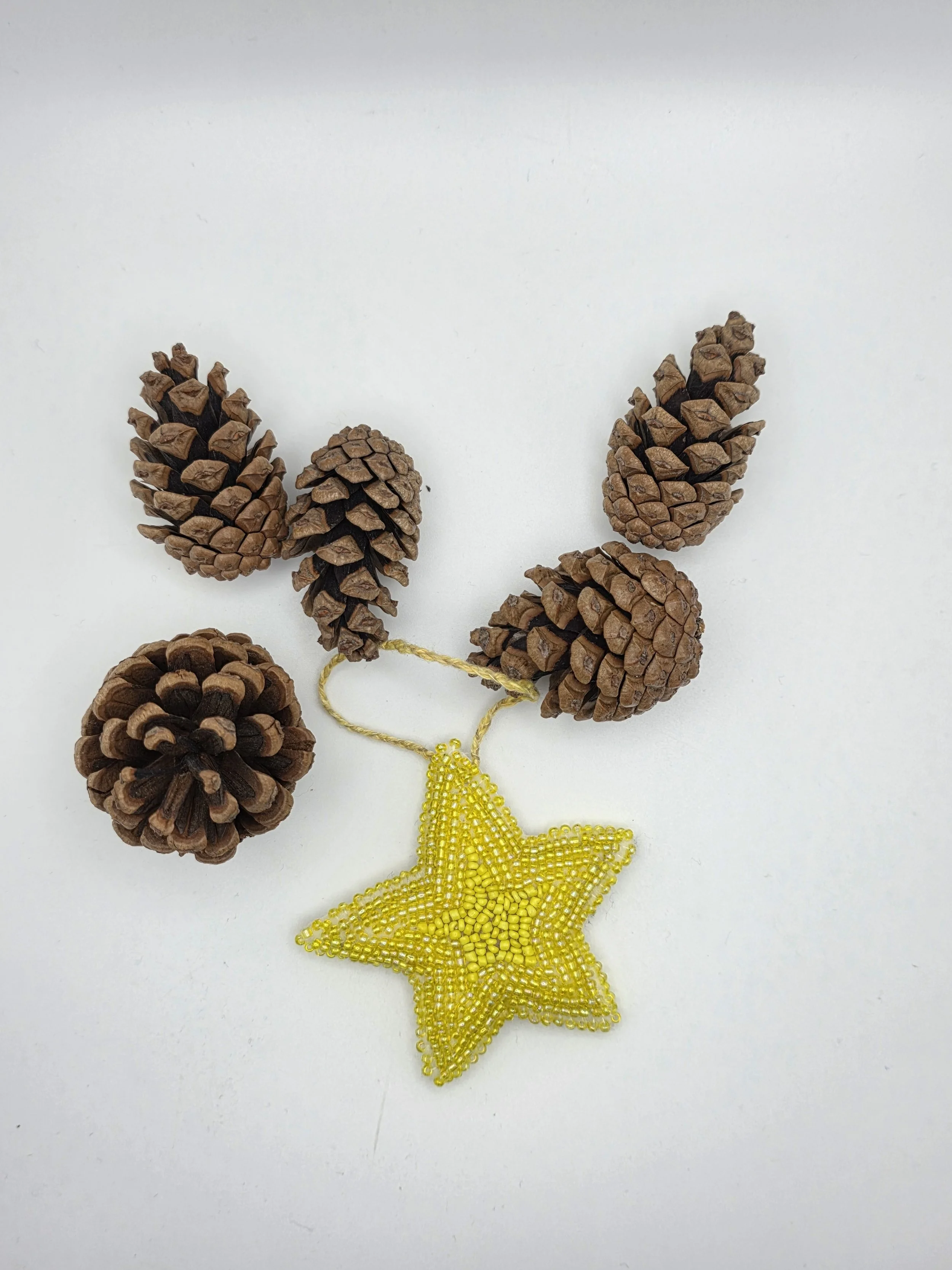 Handmade yellow beaded star decoration lying flat beside pine cones on a white background.