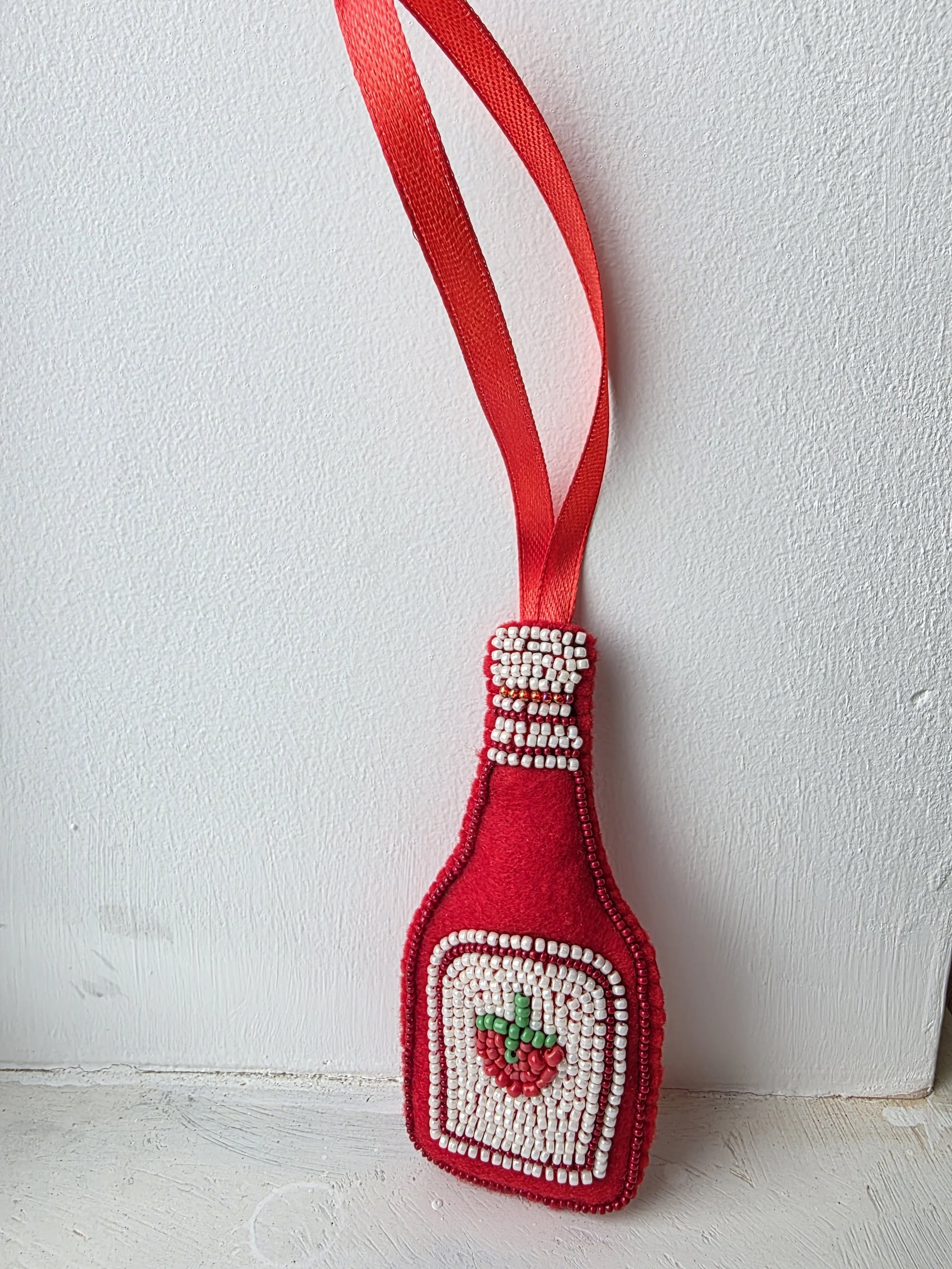 Handmade beaded tomato ketchup Christmas decoration in red felt, leaning against a white background