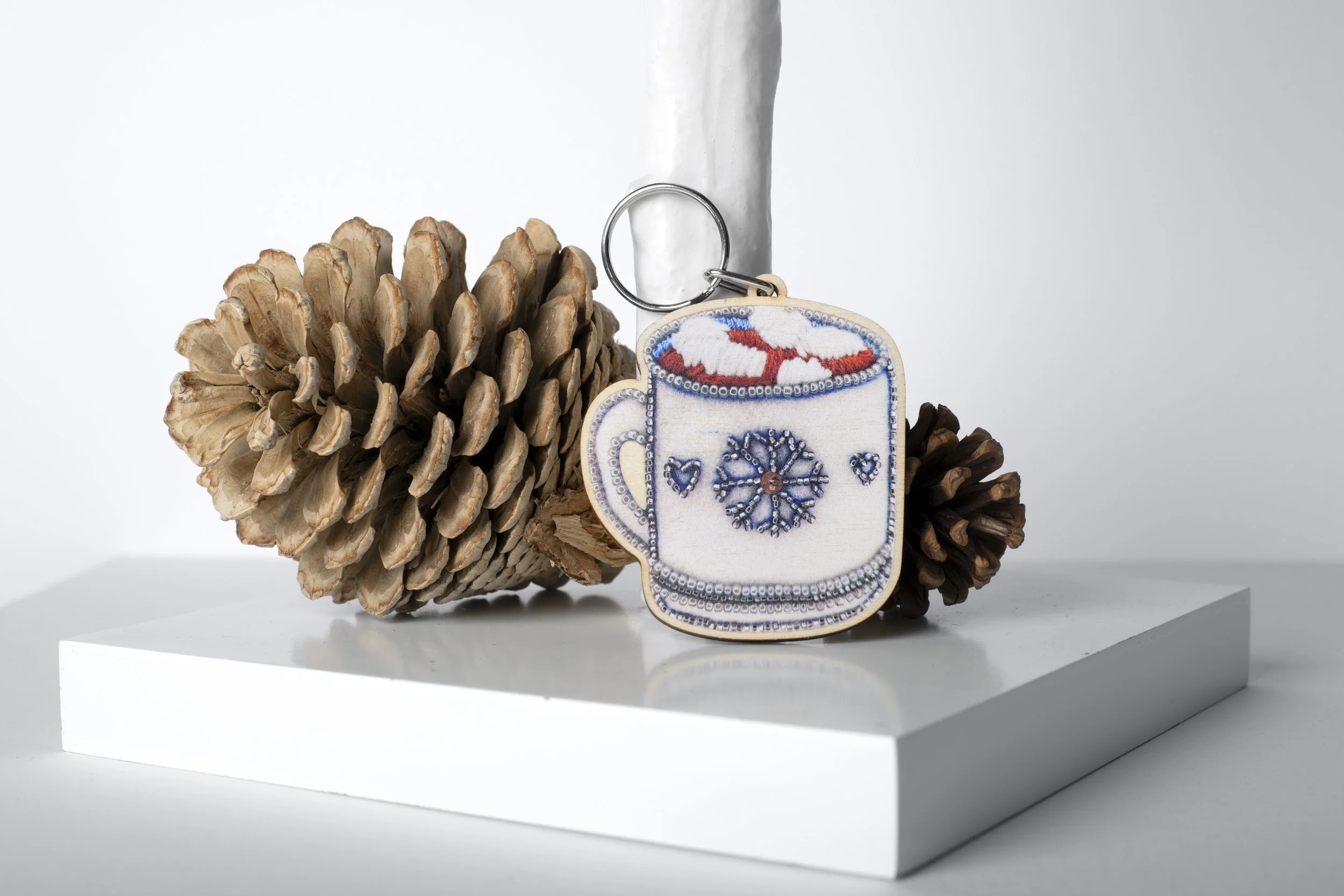 Wooden hot chocolate keyring illustrated by Hayley Mallett, displayed standing next to pine cones.