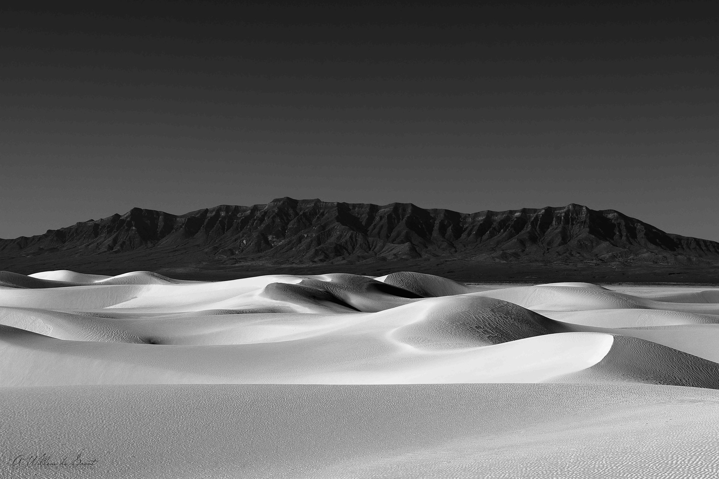 Black and white photo taken at White Sands National Park with a mountain range in the background under a clear sky.