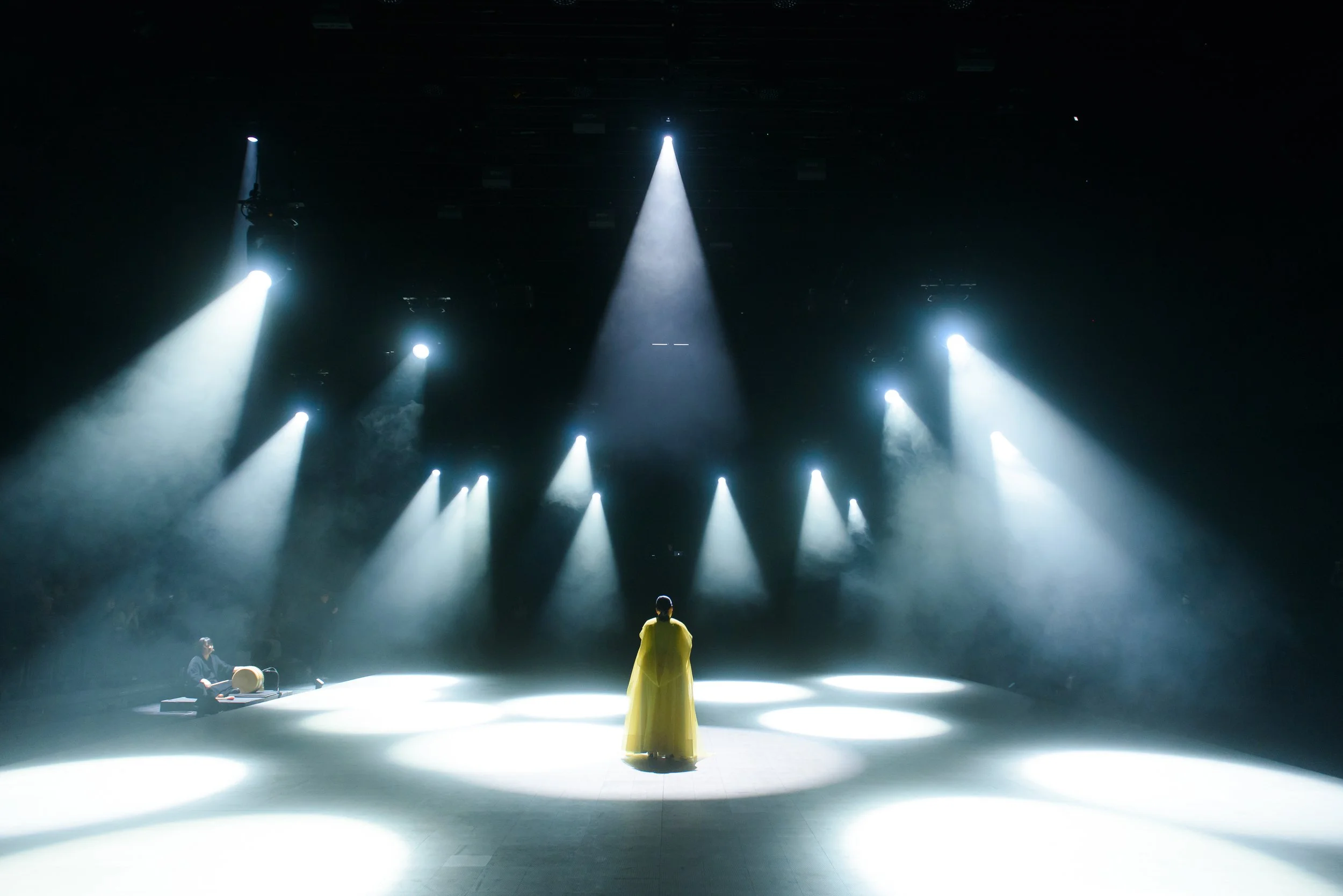 A person in a yellow dress stands on a stage under spotlight beams with smoke effects and a seated person playing a musical instrument on the side.