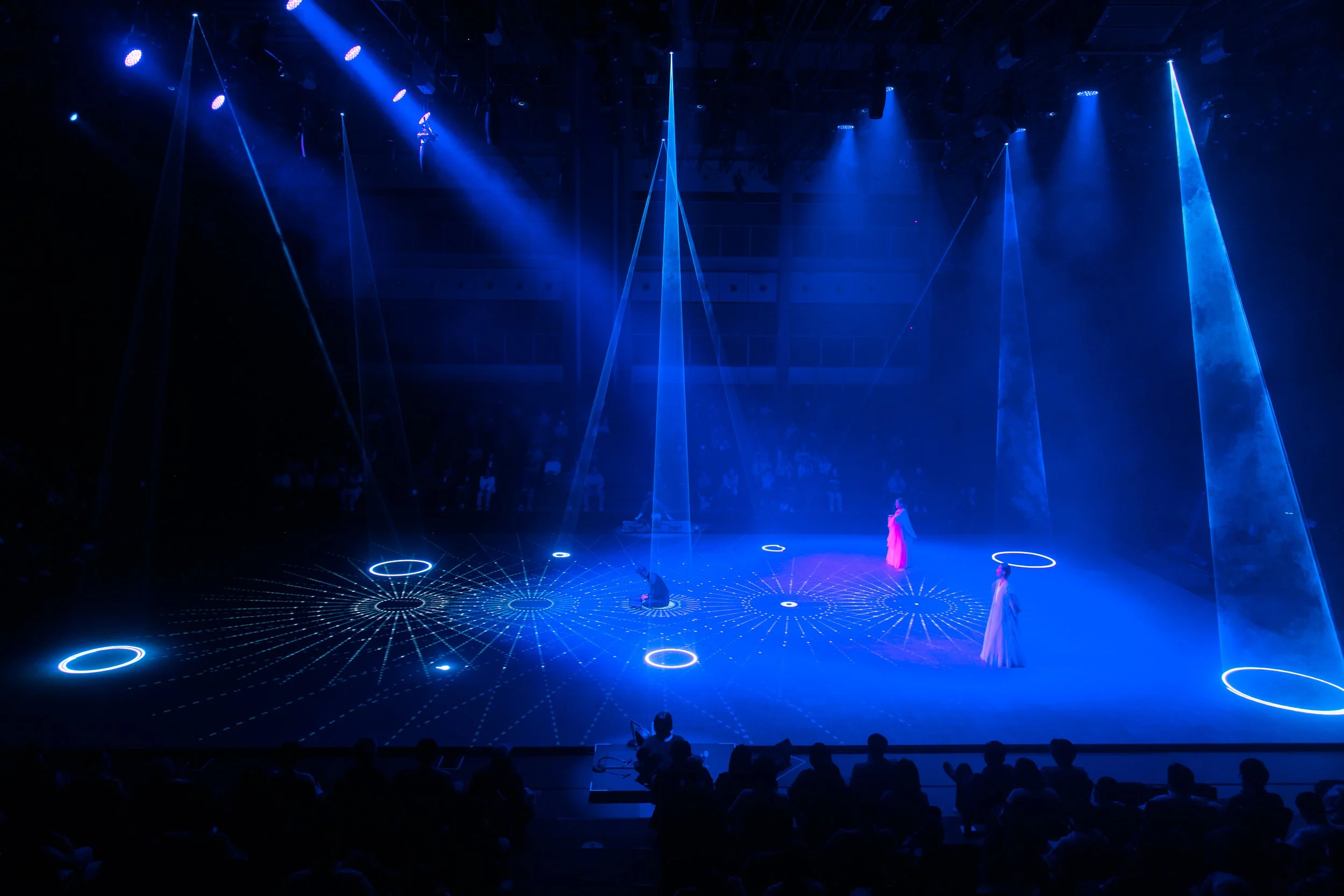 A stage performance illuminated with blue lighting and laser projections. Three performers in elegant costumes stand amidst circular and line patterns projected on the floor. Audience members are visible in the background, and beams of light highligh