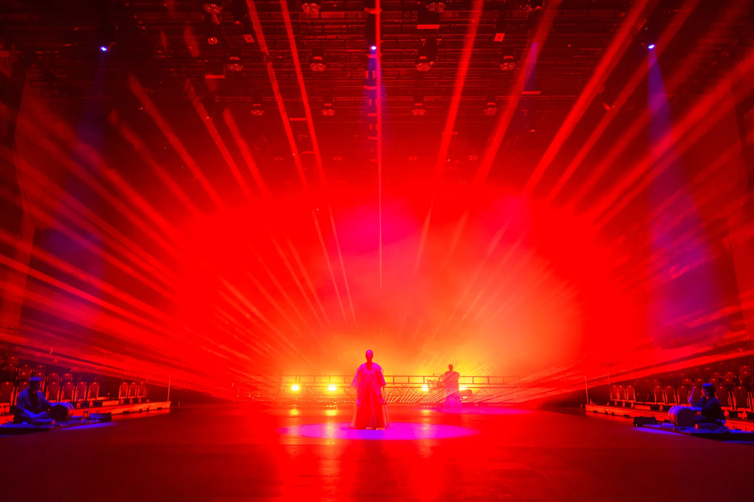 A dramatic stage performance with intense red lighting and spotlights, featuring a person standing center stage with musicians seated on either side. The background is illuminated with vibrant colors creating a dynamic visual effect.