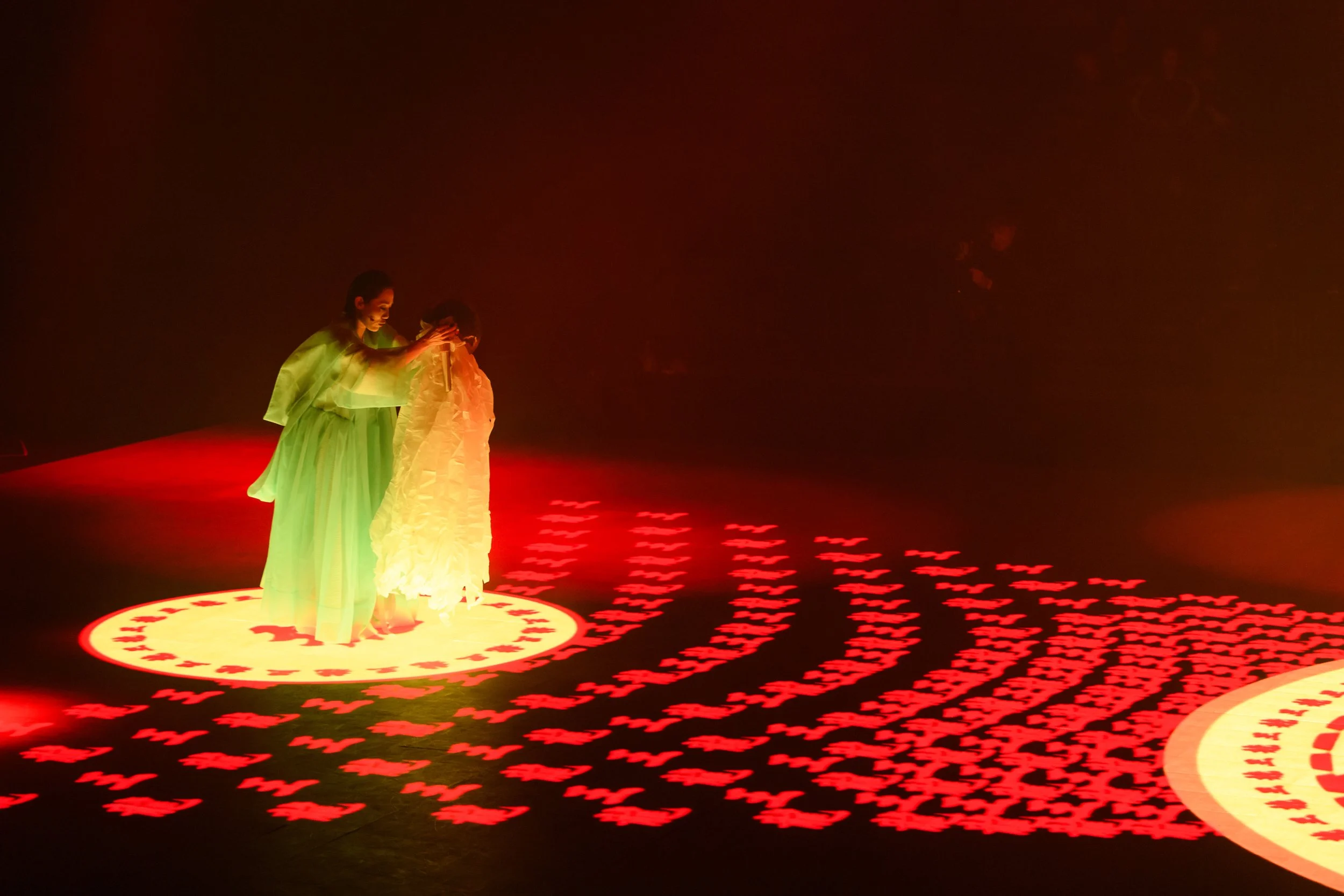 Two performers in flowing costumes under red and green stage lighting with patterned projections on the floor.