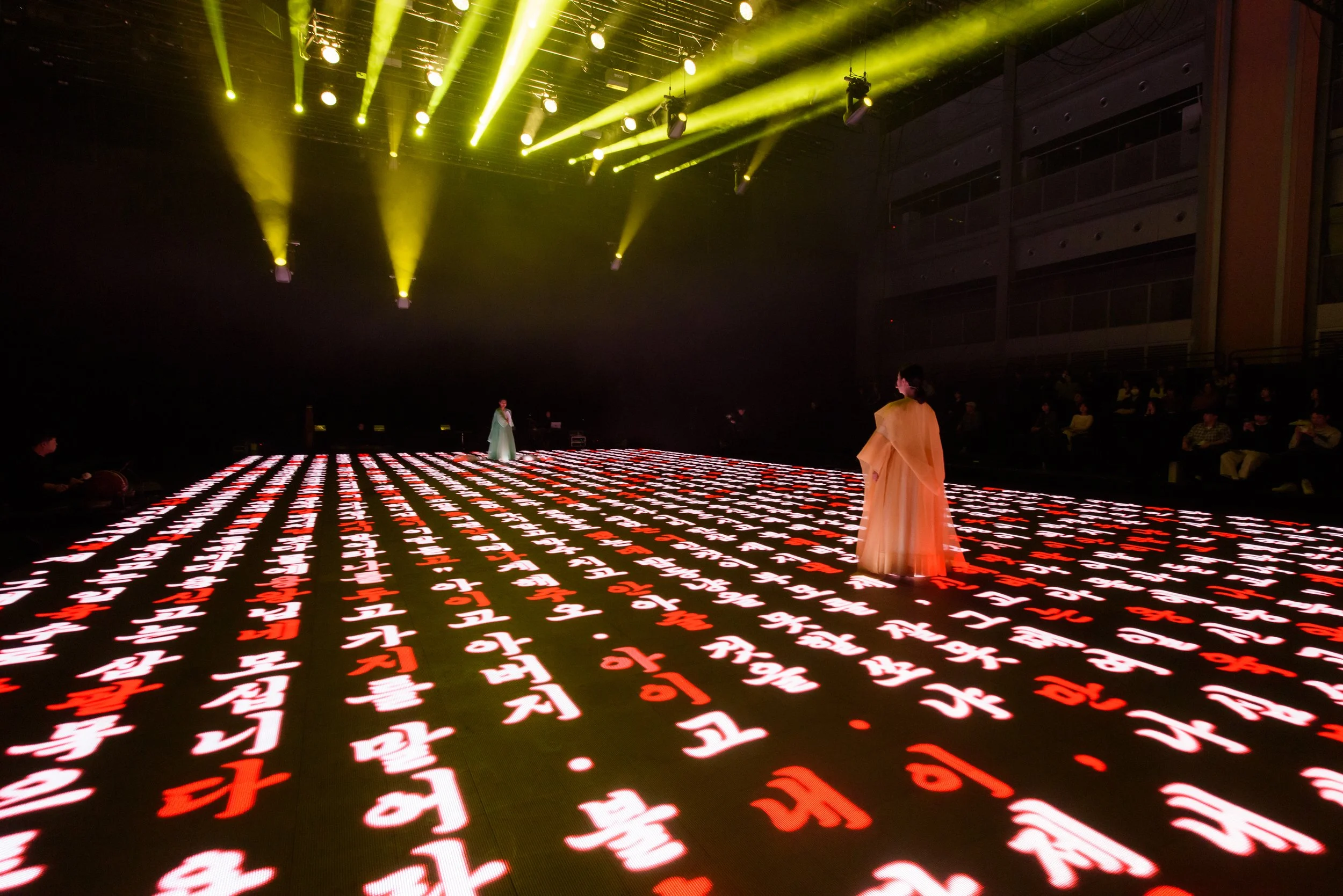 Two people in traditional attire stand on a stage with glowing red and white Korean characters projected on the floor, under a ceiling with bright yellow stage lights.