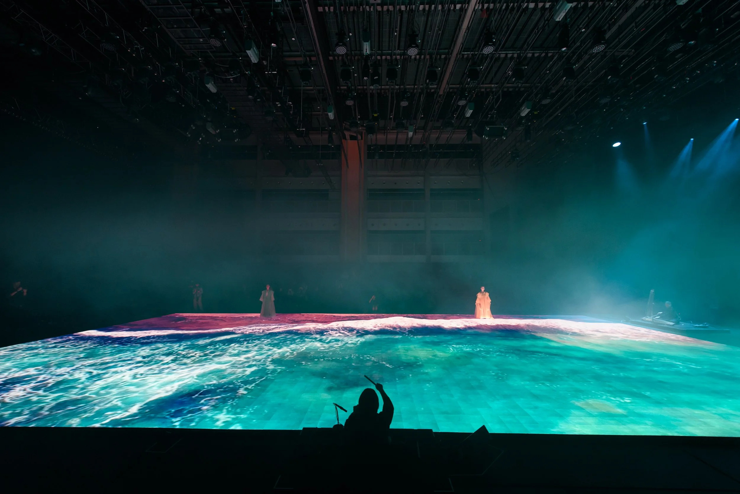 Theater performance with digital water projection on stage, overhead lights, silhouetted figure in foreground, and two people in costumes on stage.
