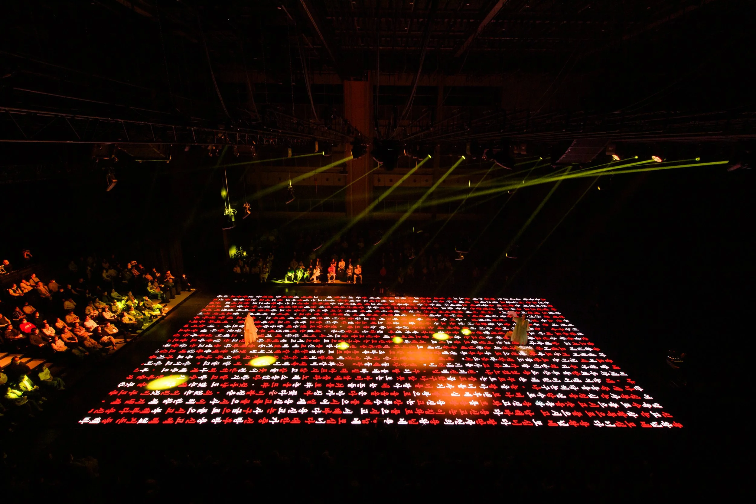 People watch a performance on a stage with illuminated red and white patterns and beams of light.
