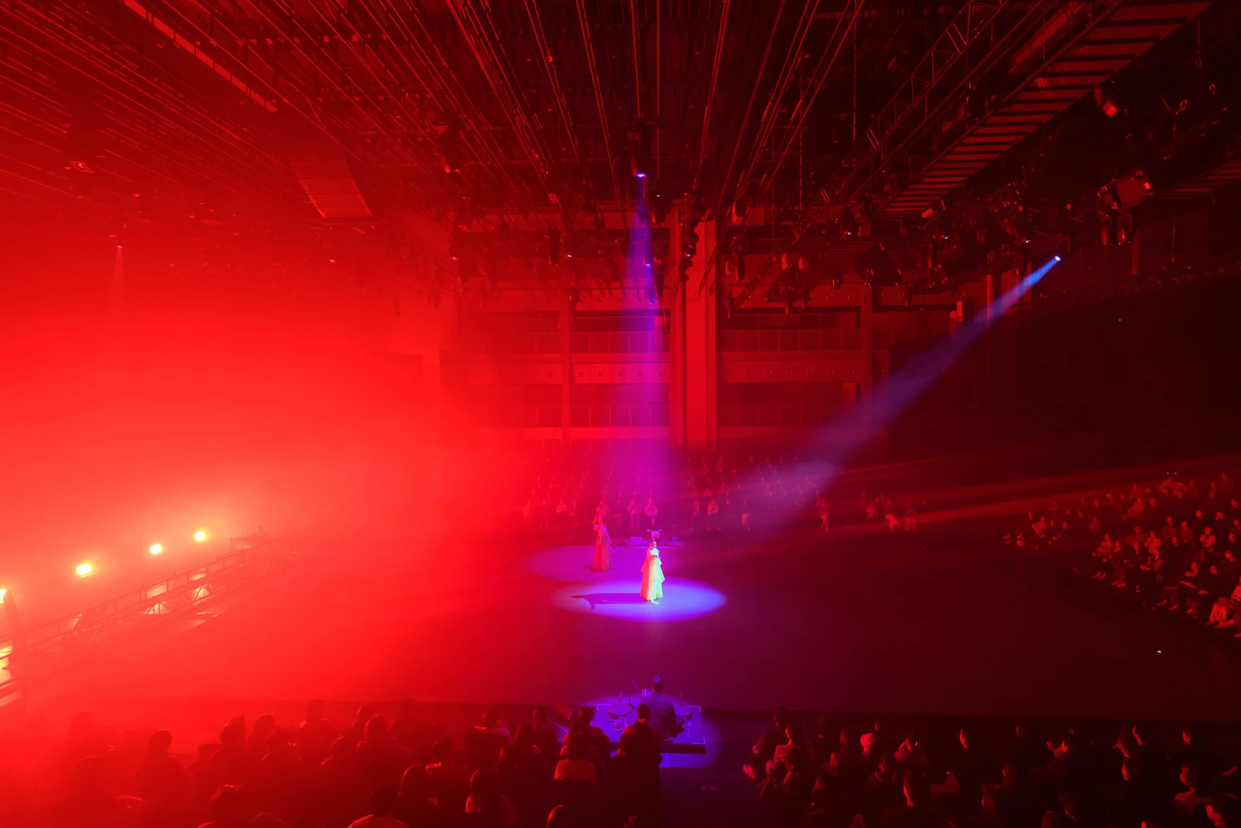 Theatrical performance with dramatic red lighting and spotlights highlighting performers on stage, viewed by a seated audience.