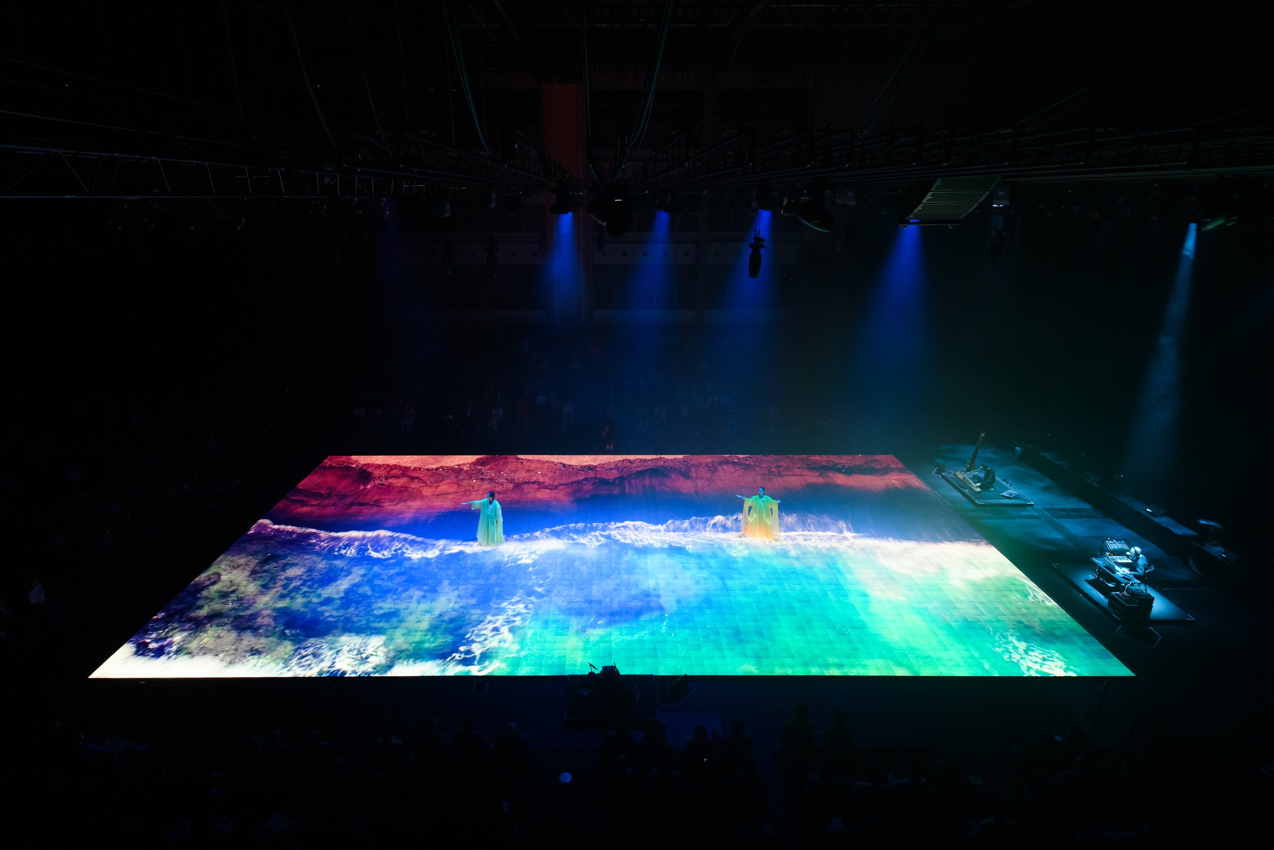 Large digital floor display with colorful ocean waves and beach scenery projected, two performers in green costumes, stage lighting, and technical equipment in a dark room.