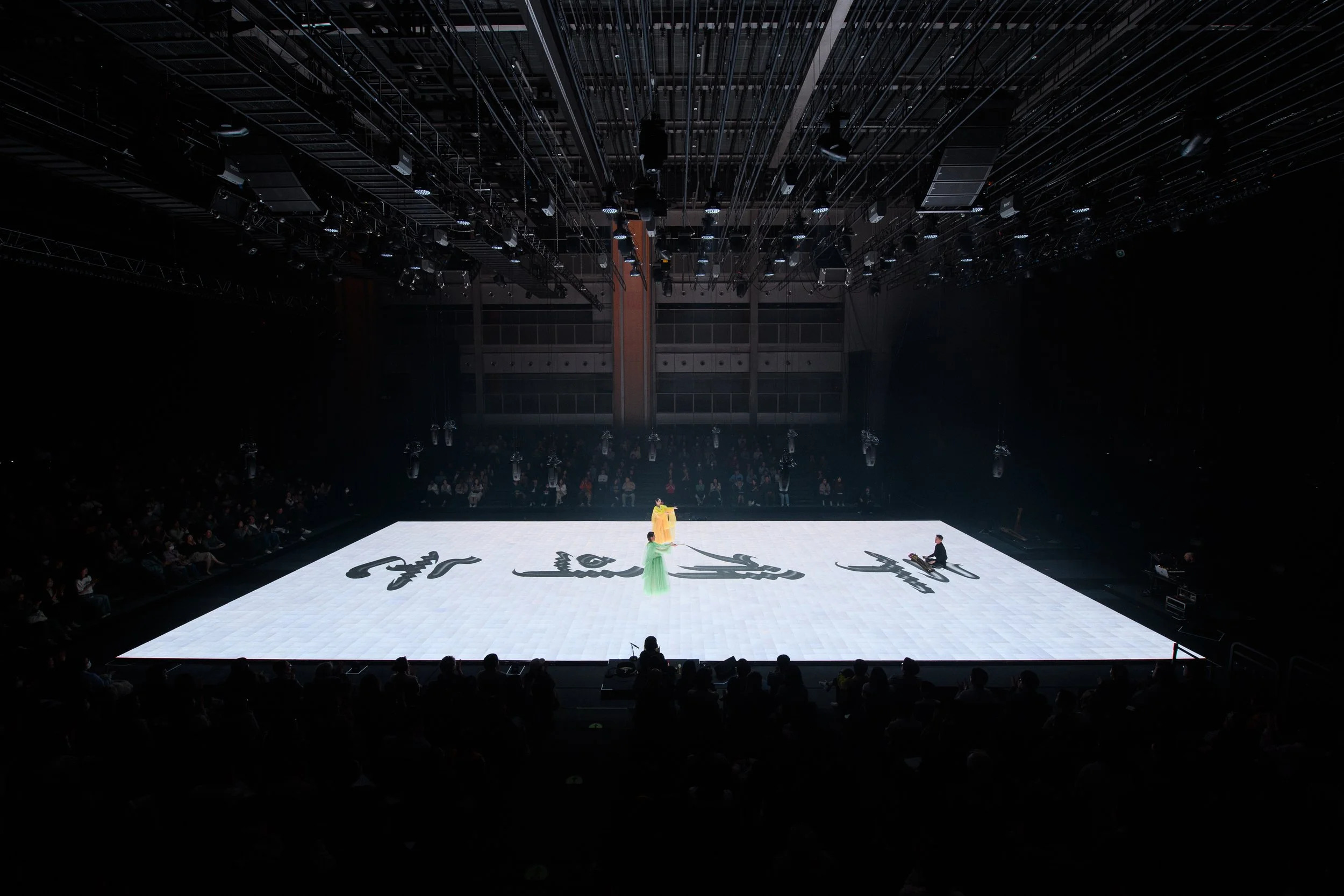 Large stage with bright white floor, featuring two performers in colorful outfits; audience seated in the background.