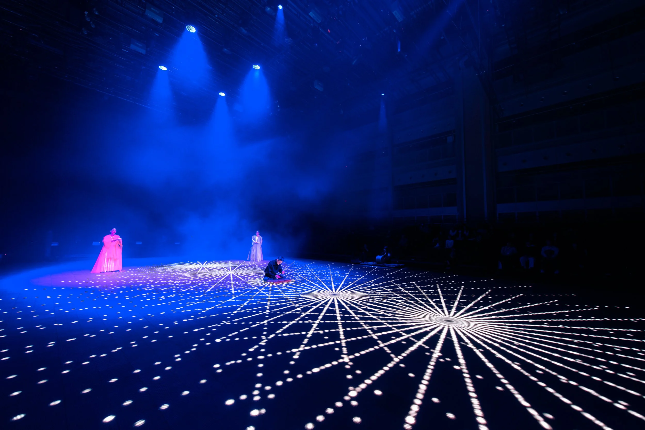 Stage performance with blue lighting and geometric light patterns on the floor, featuring three performers, one kneeling and two standing.
