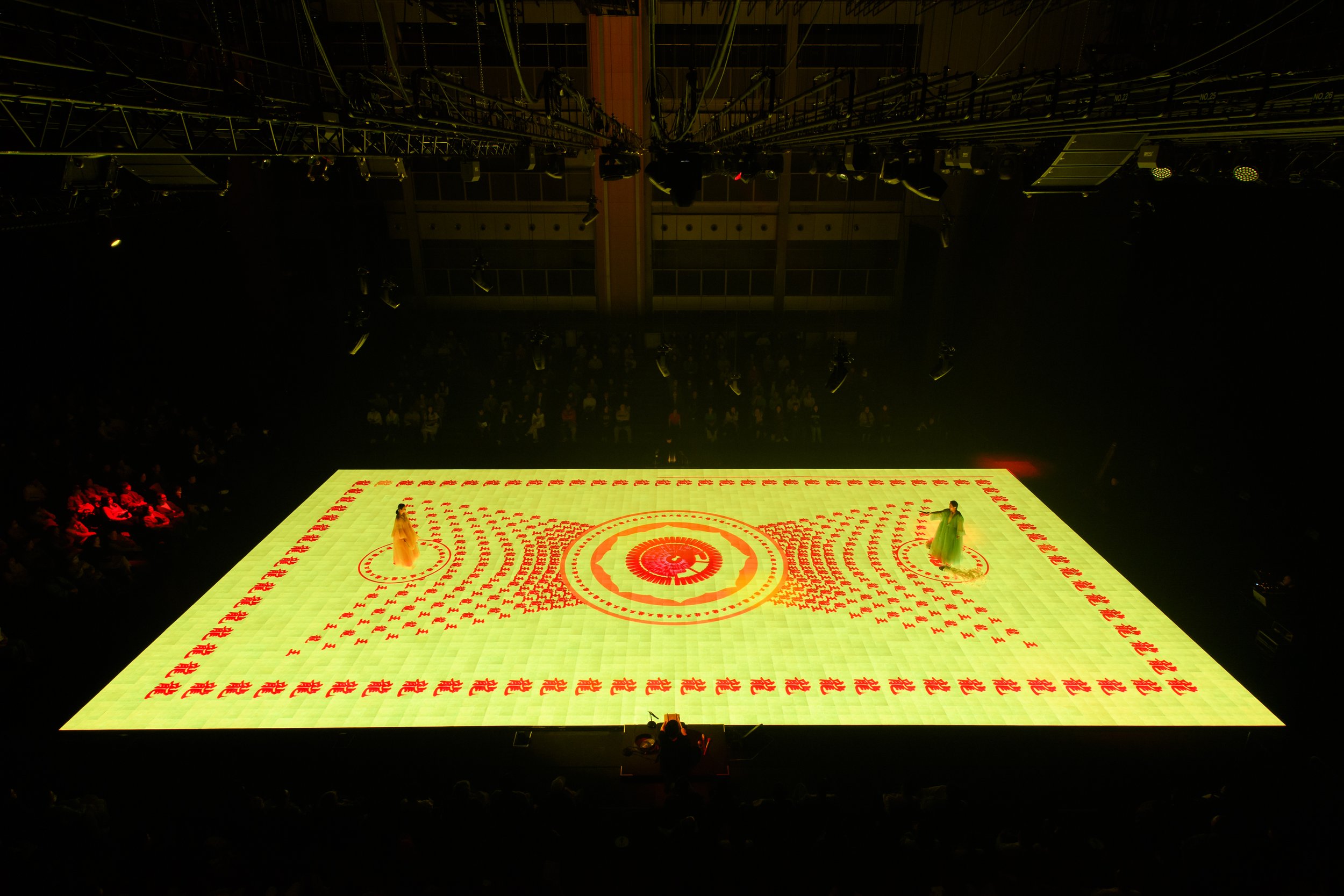 Stage with intricate light pattern, featuring two performers in costumes, surrounded by an audience and stage equipment above.