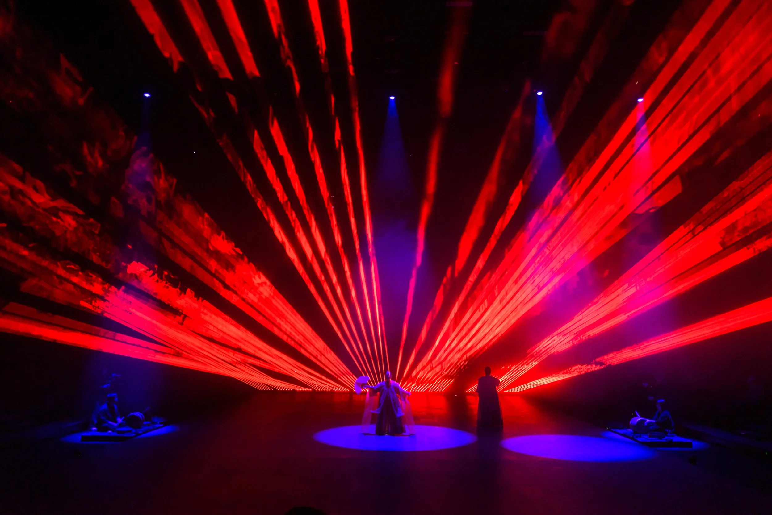 A vibrant stage performance with red laser lights fan out behind silhouetted performers."}